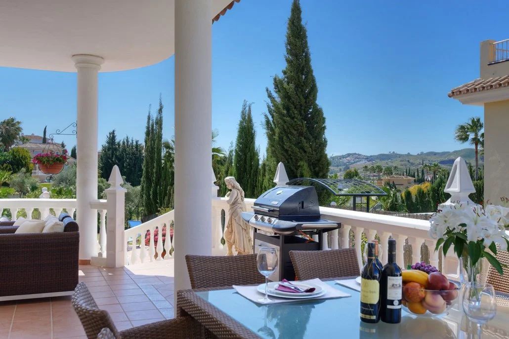 Outdoor patio with dining table set for dinner, barbecue grill, lush green trees, and a scenic hillside view in the background.