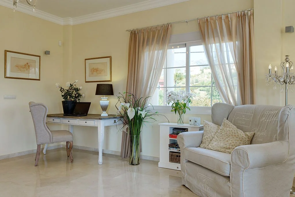 Bright living room with cream-colored walls, a window with beige curtains, a white armchair with pillows, a small white side table with books, a large floral vase, a desk with a lamp and laptop, and framed art on the walls.