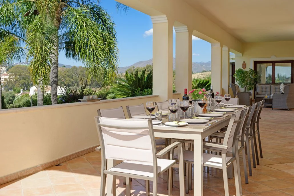 An outdoor dining area with a long table set with wine glasses, plates, and utensils, overlooking lush greenery and mountains in the background.