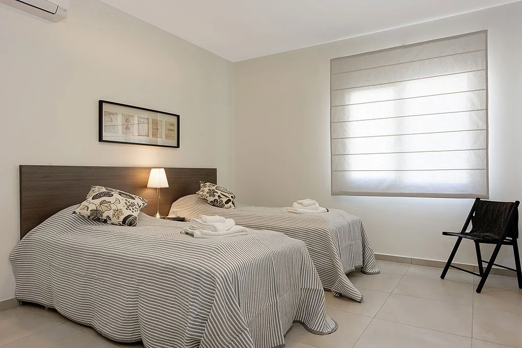 A bedroom with two twin beds, each with striped bedspreads and decorative pillows, a wall-mounted picture above the beds, a small lamp between the beds, folded towels on each bed, a black chair near a large window with a soft shade, and a white wall.