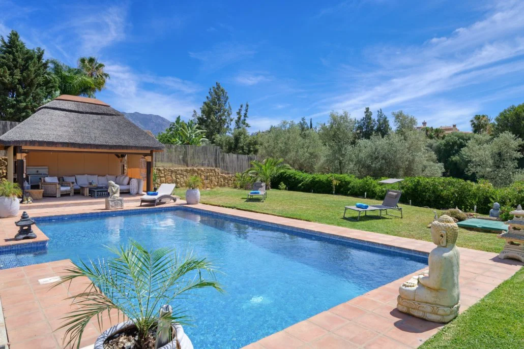 A backyard swimming pool area with lounge chairs, a thatched-roof gazebo, tropical plants, statues, and a manicured lawn under a blue sky.
