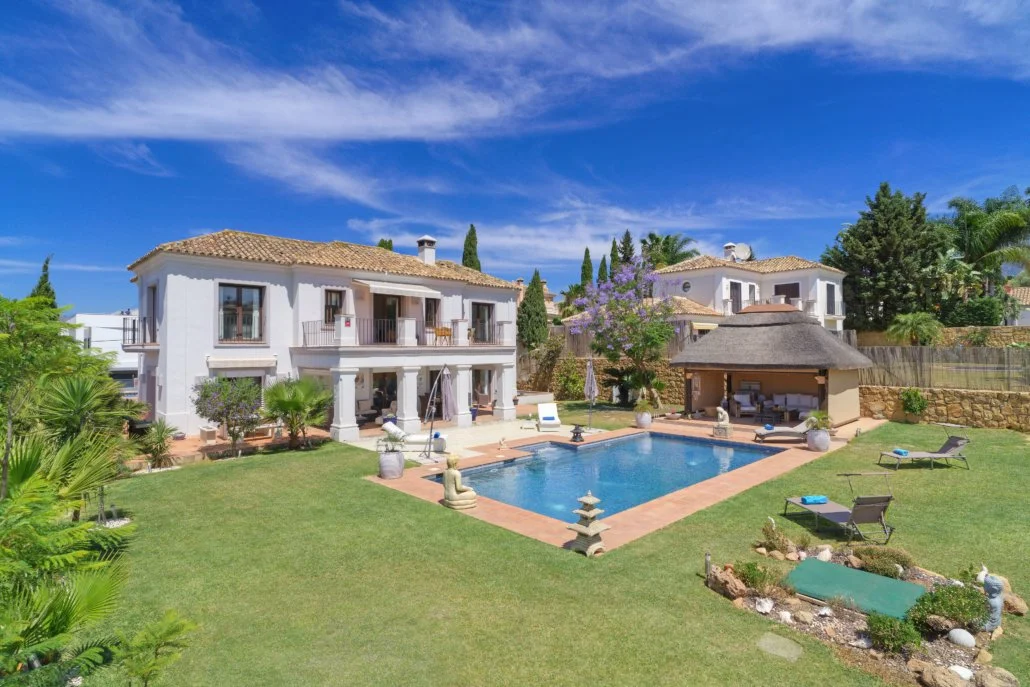 A luxurious backyard with a swimming pool, surrounded by a lush green lawn, palm trees, a two-story white house with balconies, and a tiki hut with outdoor furniture under a blue sky with wispy clouds.