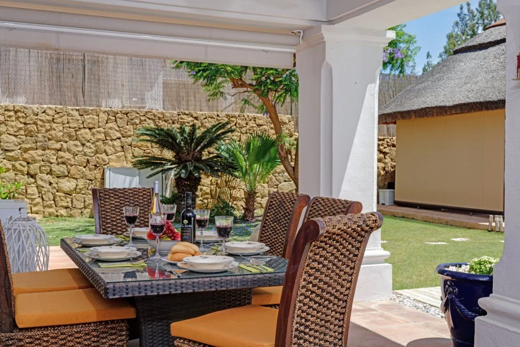 Outdoor dining table set with wine, grapes, bread, and glasses on patio, with trees and stone wall in background.