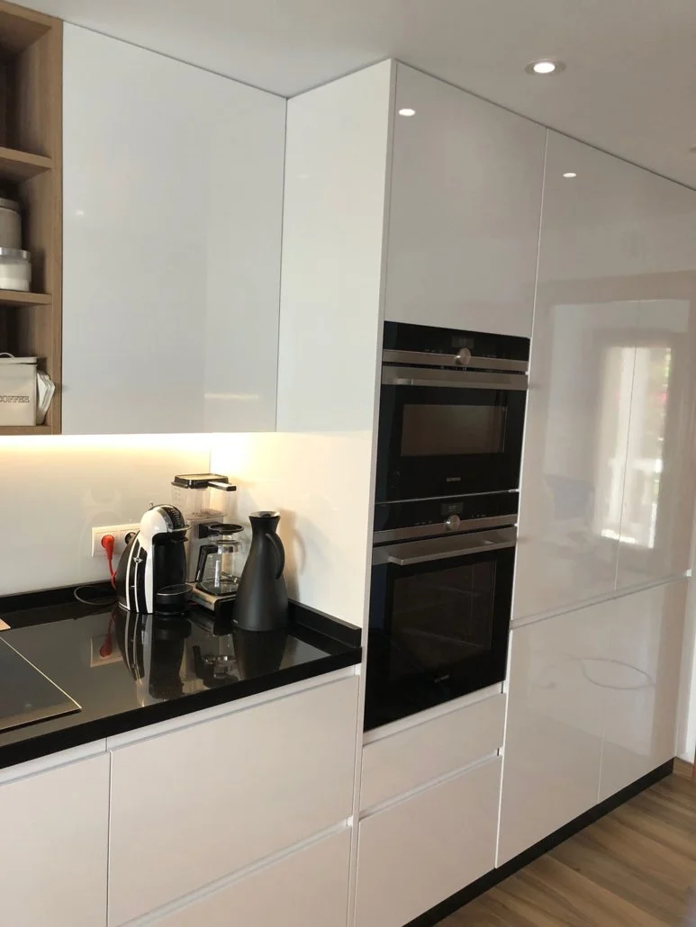 Modern kitchen with white cabinetry, black countertops, built-in oven and microwave, and small appliances including a tea kettle, coffee maker, and grinder on the counter.