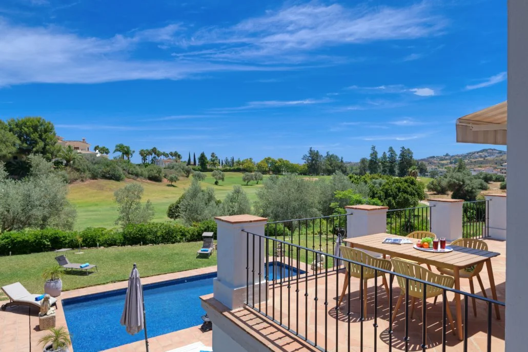 Backyard patio view with table, chairs, umbrella, pool, green grass, and scenic hillside with trees and houses under a blue sky.