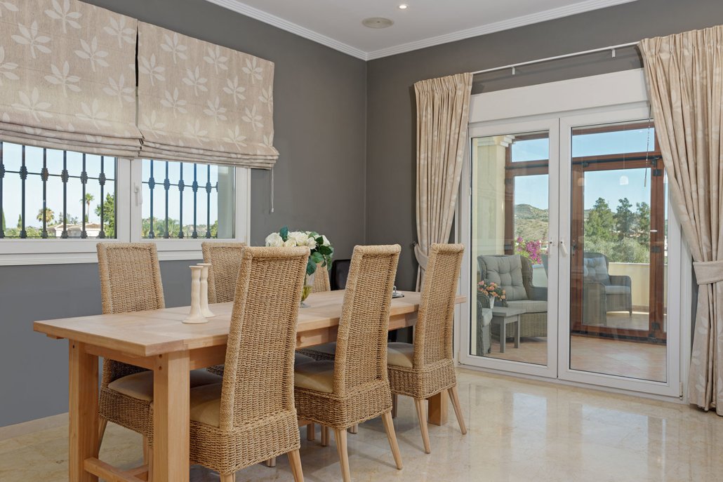 Dining room with wooden table, six wicker chairs, and large glass sliding door leading to outdoor patio with seating and trees.
