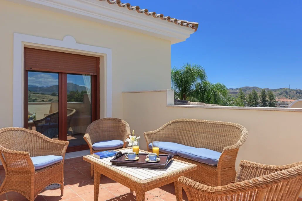A sunny outdoor balcony with wicker furniture including a loveseat, two chairs, and a table with breakfast items. The table has two glasses of orange juice, two cups of coffee, and a small flower arrangement. There is a sliding glass door with a wood
