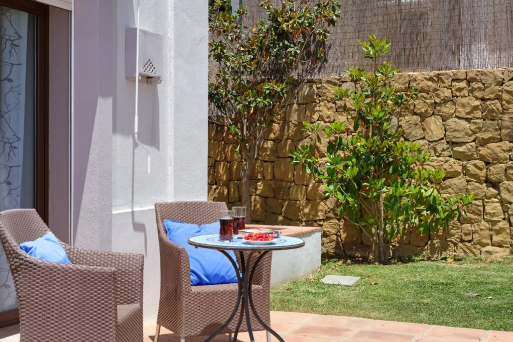 Patio with two wicker chairs with blue cushions, a small round table with glasses of red drink and a plate of strawberries, green grass, a stone wall, and a tree in a backyard.