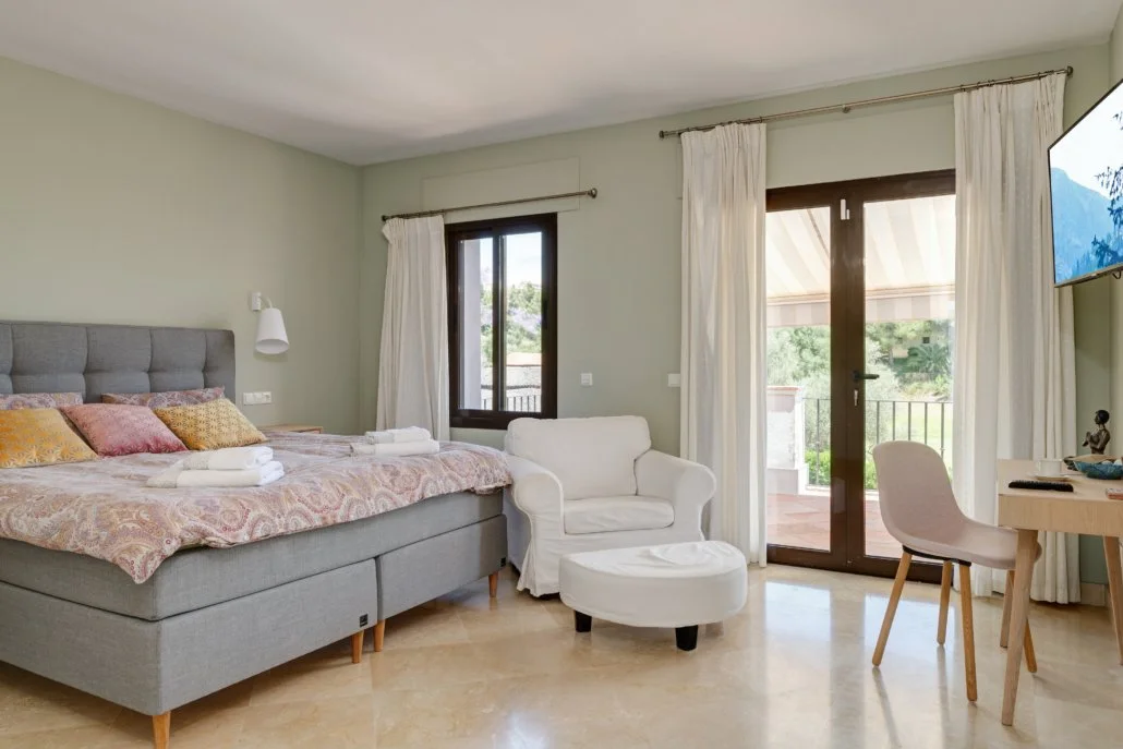 A hotel room with a large bed, an armchair with an ottoman, a small desk with a chair, and a balcony with glass doors. The room has light-colored walls, curtains, and wooden flooring.