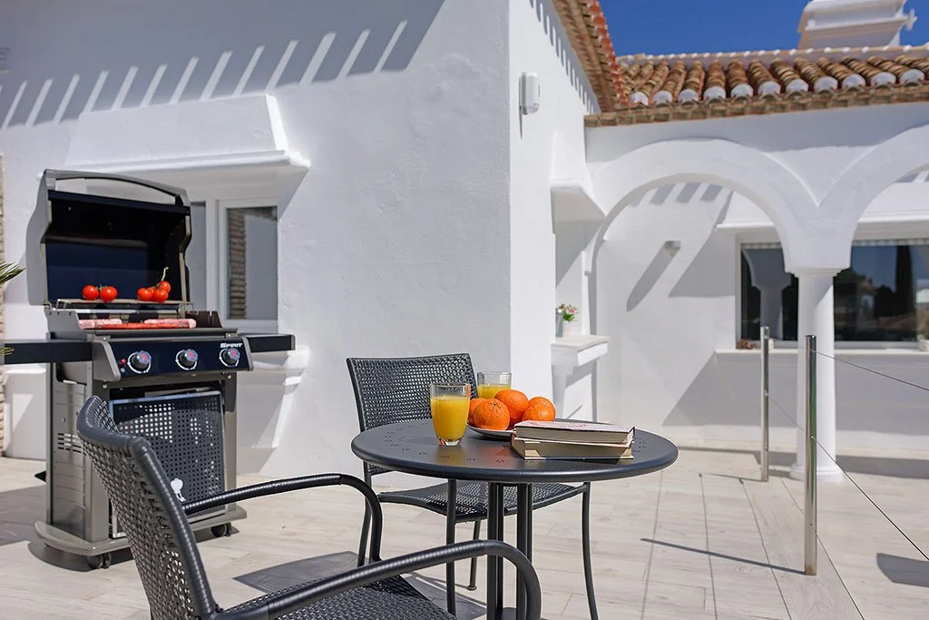 Outdoor patio with a barbecue grill, a round table with two glasses of orange juice, a plate of oranges, books, and black chairs, with white stucco building and tiled roof in the background under a blue sky.