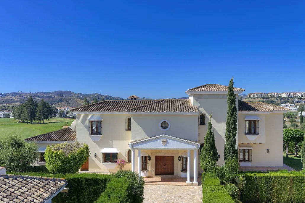 Large Mediterranean-style house with white stucco walls, brown tile roof, surrounded by green trees and bushes, set against a backdrop of hills and a clear blue sky.