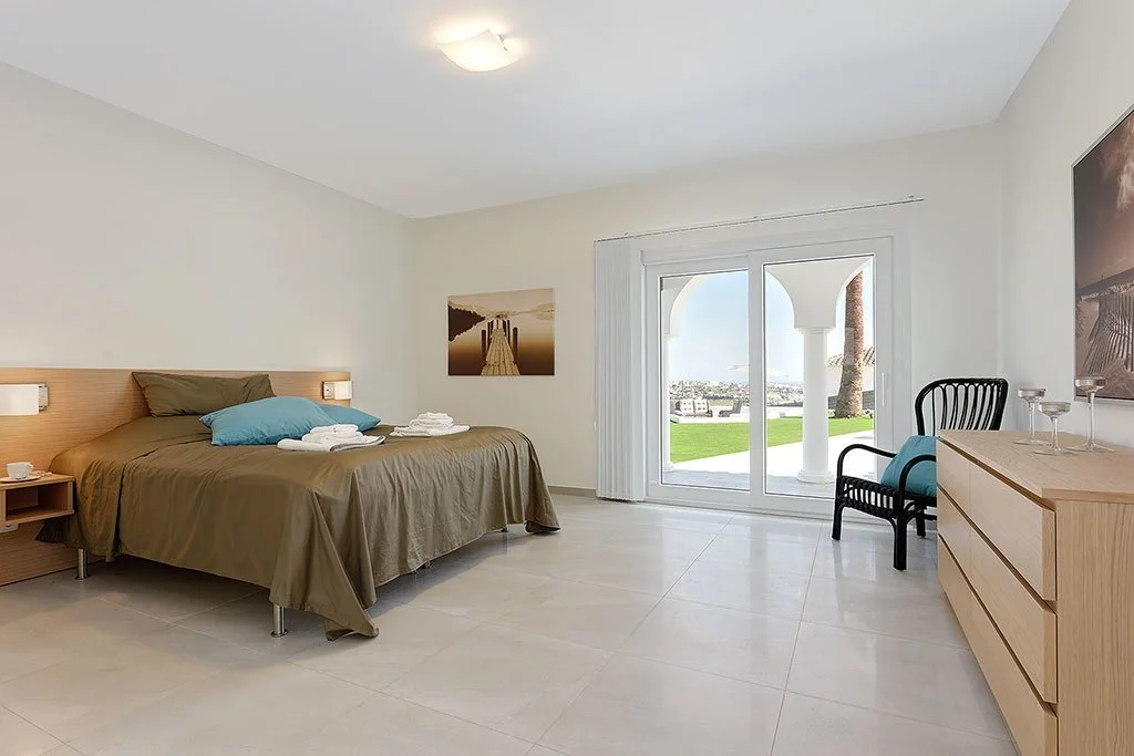 A bright, minimalist hotel room with a large bed, two pillows, and neatly folded towels. There's a nightstand with a small cup, a seating area near a sliding glass door, and a scenic view outside with grass and columns.
