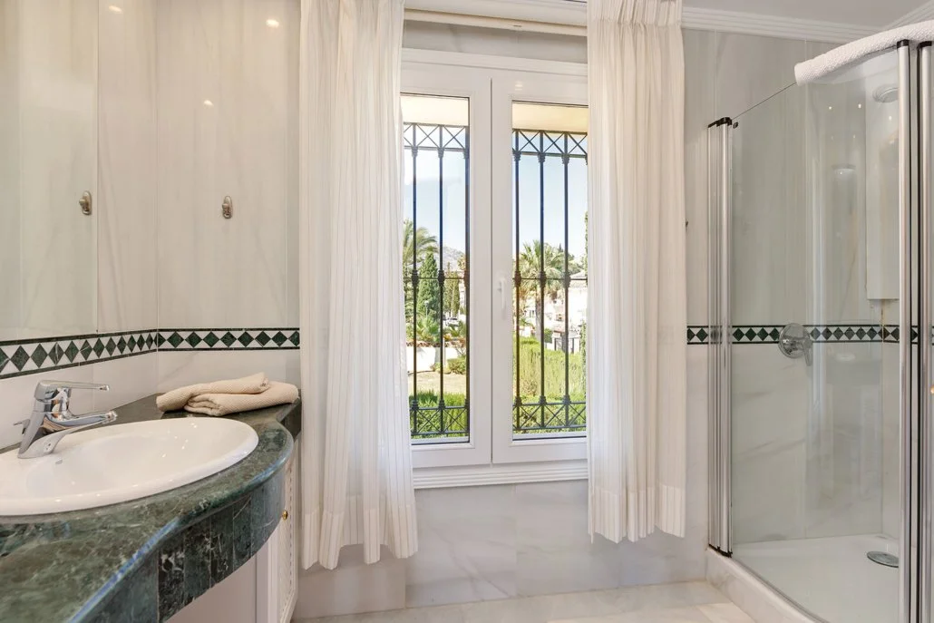 View of a bathroom with a window showing outdoor greenery, a shower stall, a countertop with a sink, and white curtains.