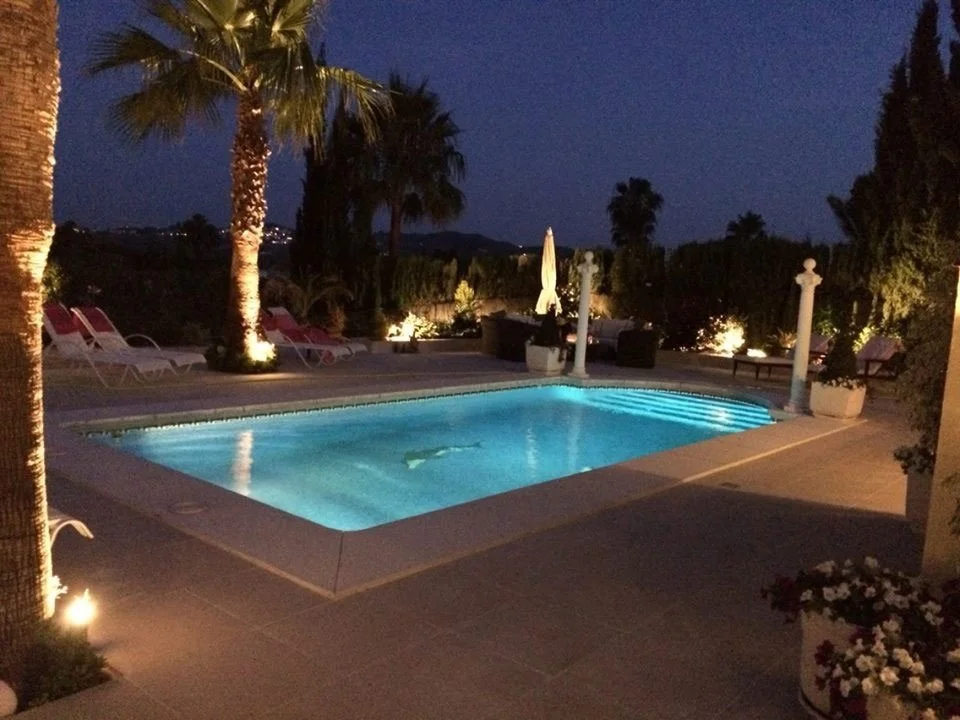 A backyard pool illuminated at night, surrounded by palm trees, lounge chairs, and outdoor furniture with decorative lighting.