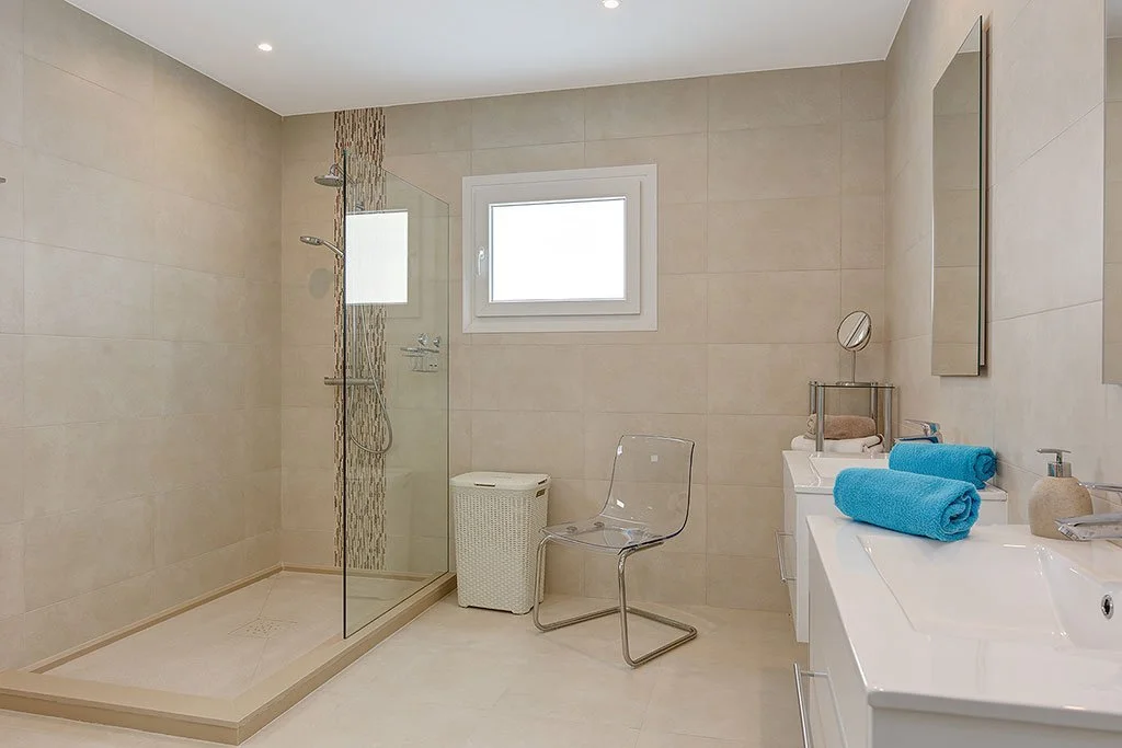 Modern bathroom with walk-in shower, small window, transparent chair, laundry basket, towels, and bathroom fixtures.