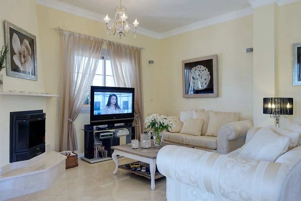 Living room with cream-colored sofas, a television on a stand, a coffee table with candles and flowers, a chandelier, wall art, a fireplace, and curtains covering a window.