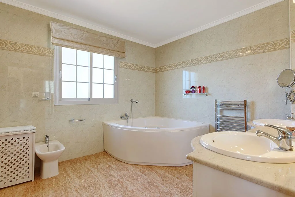 A spacious bathroom with a window, a bathtub, two sinks, a bidet, a towel rack, and decorative wall tiles.