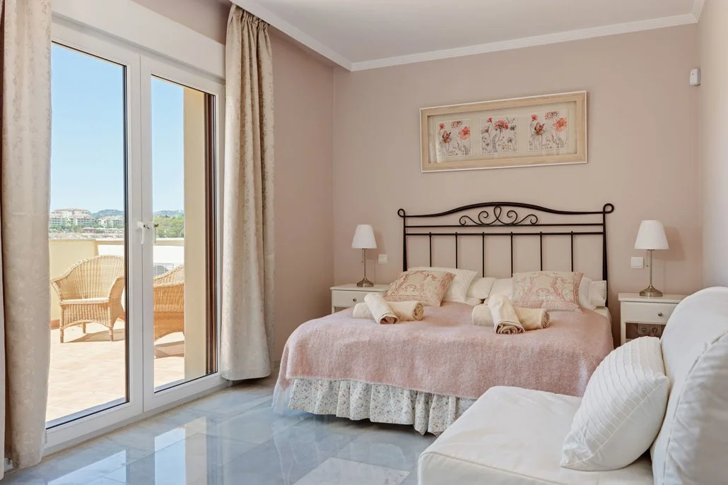 Bedroom with a metal bed frame, pink bedspread, two white nightstands with lamps, framed floral artwork above the bed, and glass door leading to a balcony with wicker chairs and a city view.