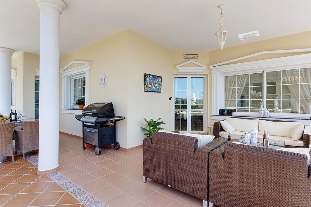 Patio area with wicker furniture, potted plants, a grill, and decorative lanterns, enclosed by a house with large windows and a sliding glass door.