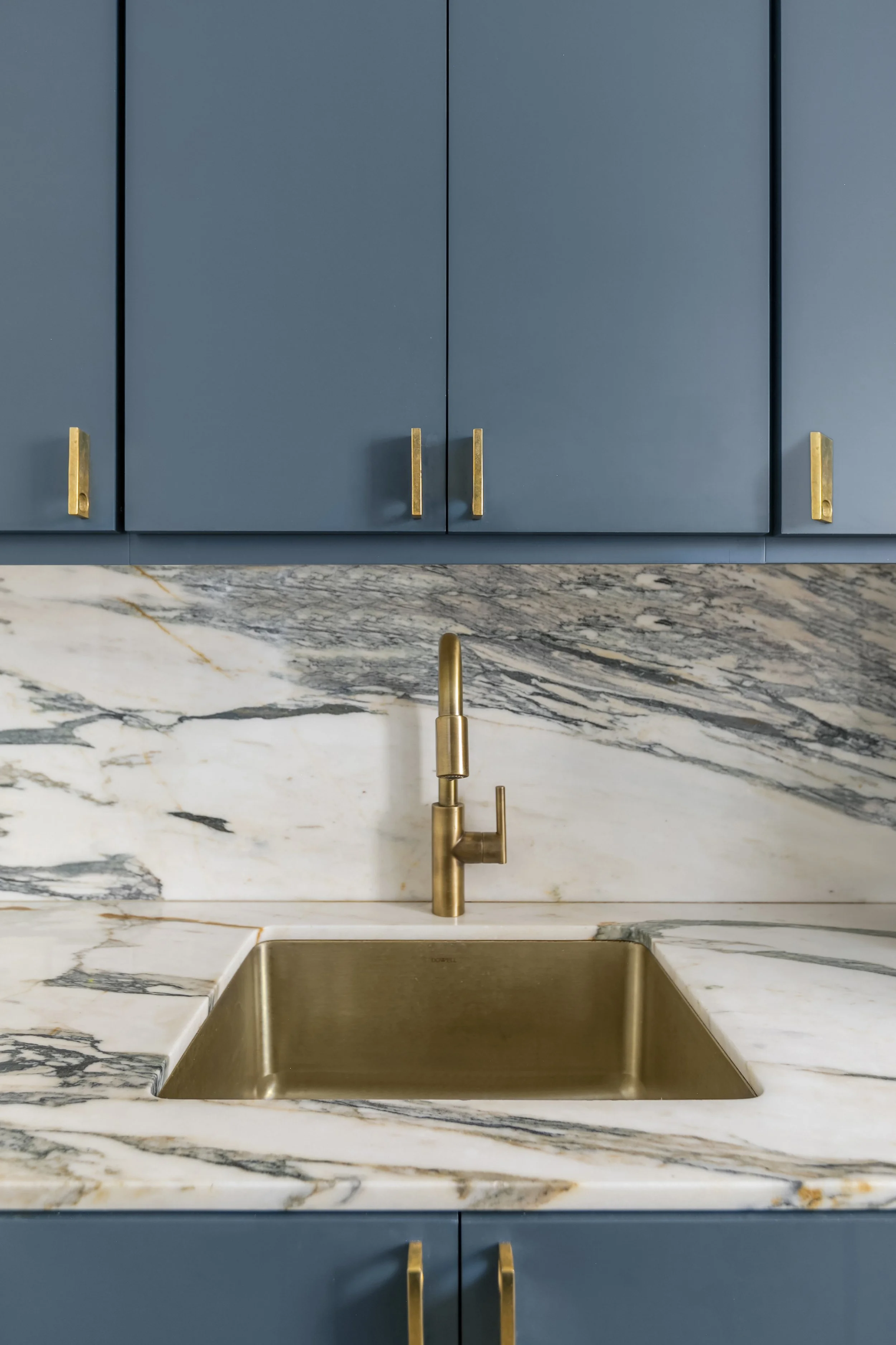 Close-up of a kitchen sink with a gold faucet, blue cabinets with gold handles, and a marble backsplash.