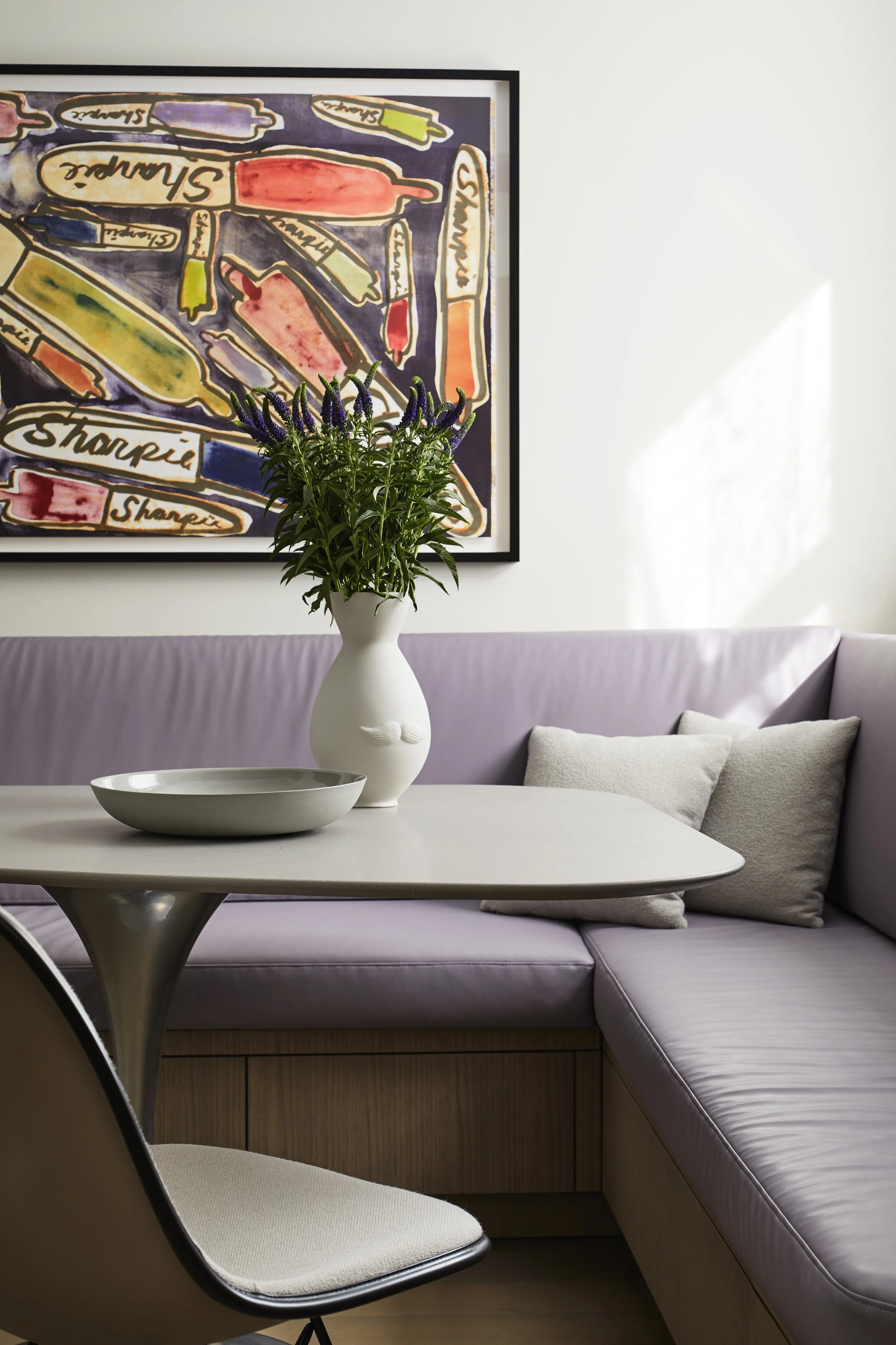 A living room corner with a white table, a lavender corner sofa with pillows, a vase with purple flowers, a decorative bowl, and colorful abstract artwork on the wall.