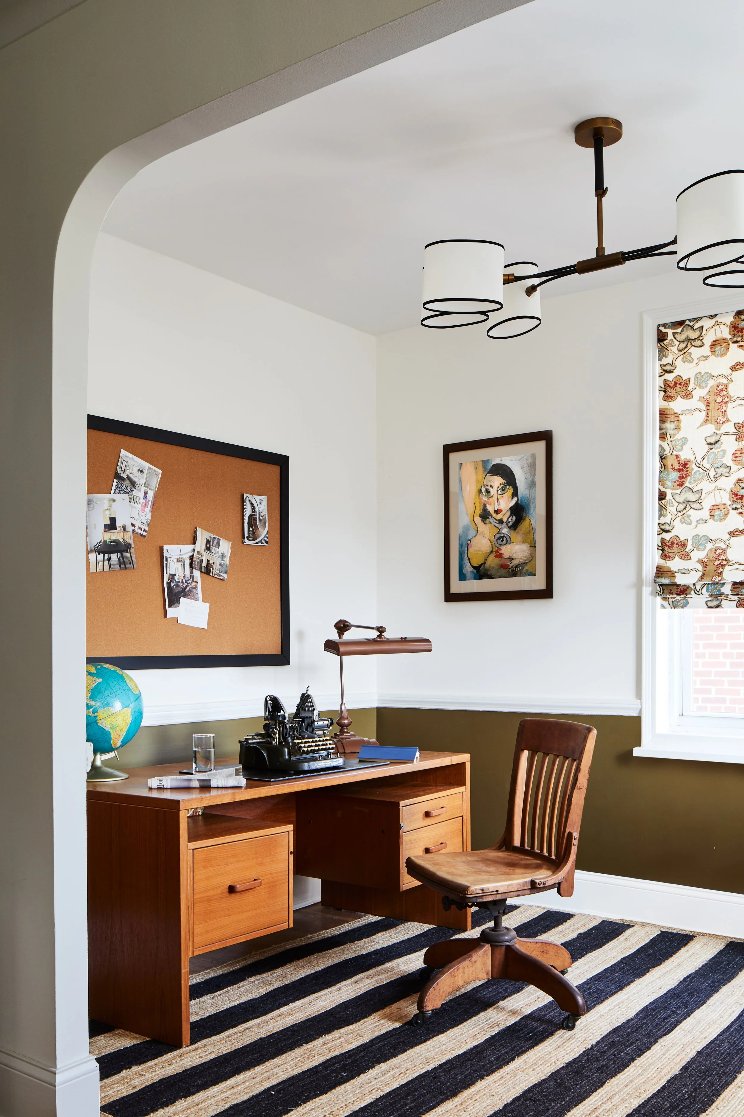 A home office with a wooden desk, vintage typewriter, globe, desk lamp, and a glass of water, set against white walls with a picture and pinboard, a window with patterned blinds, and a striped rug.