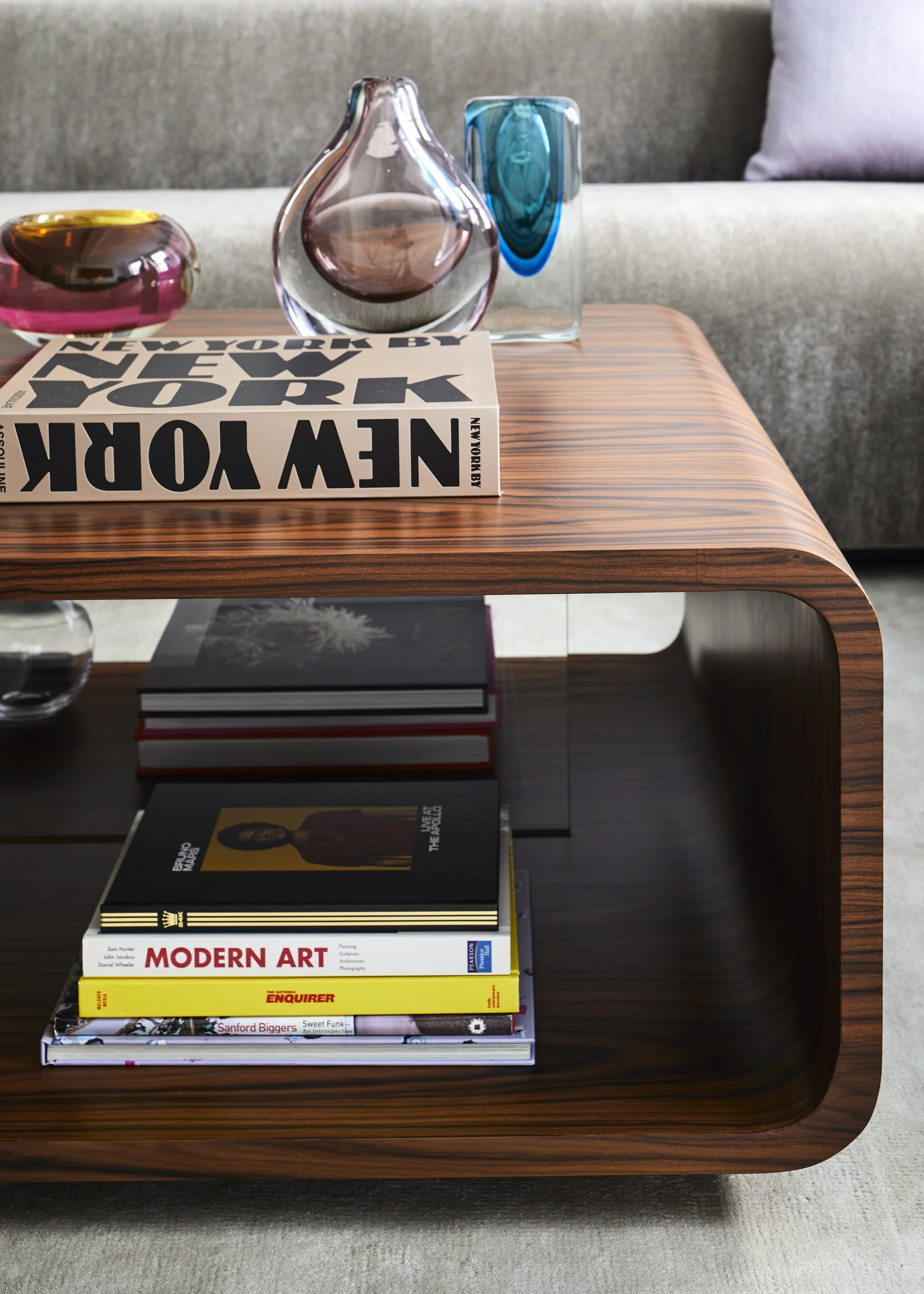 A wooden coffee table with a stack of art and design books on the shelf below, a magazine titled 'New York' on top, and three decorative glass vases on the table surface. The background features a gray sofa and a pink cushion.