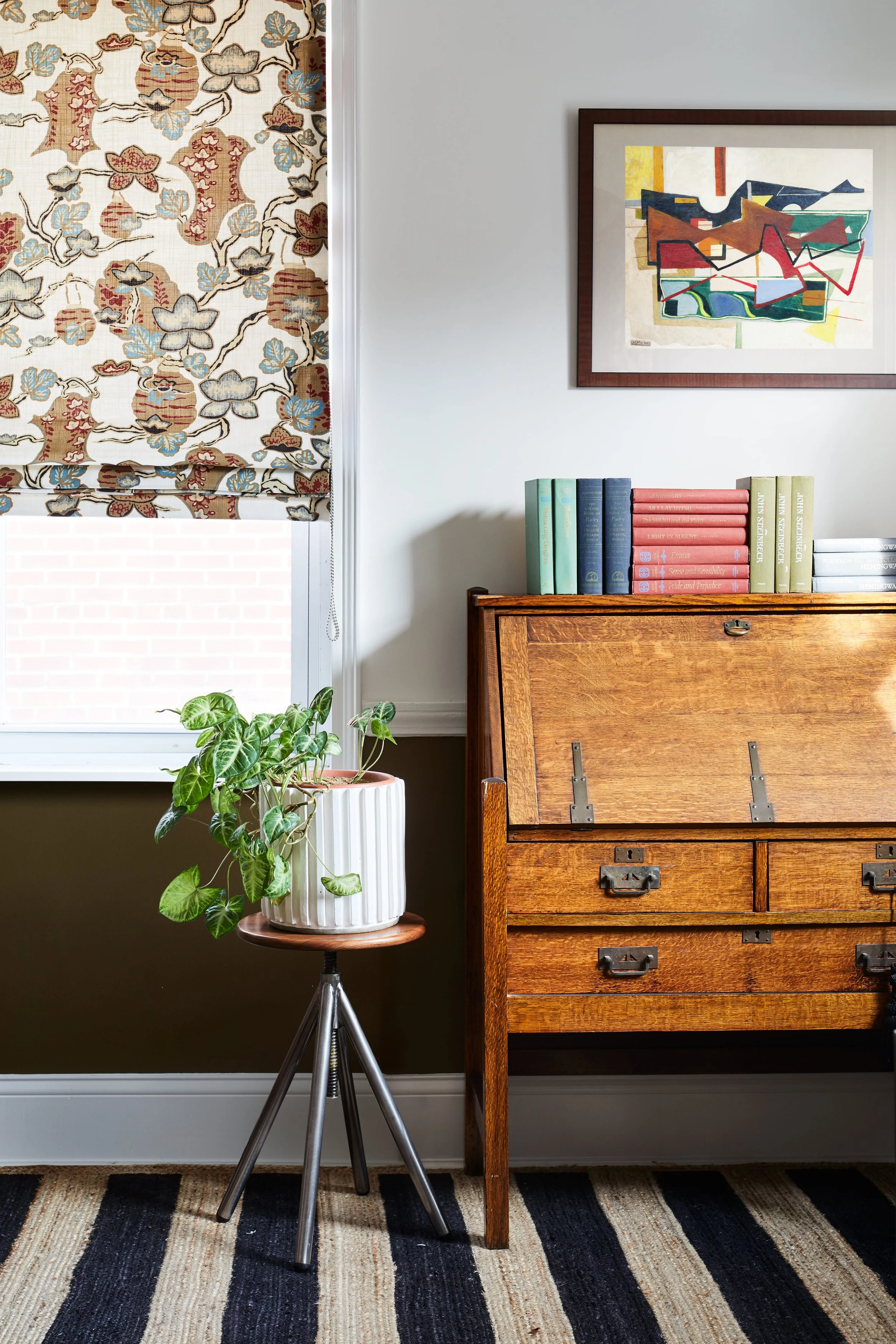 A living room corner with a window covered by a patterned curtain, a potted plant on a tripod stand, a wooden writing desk with drawers, and a bookshelf with colorful books. There's an abstract painting hanging on the wall above the desk.