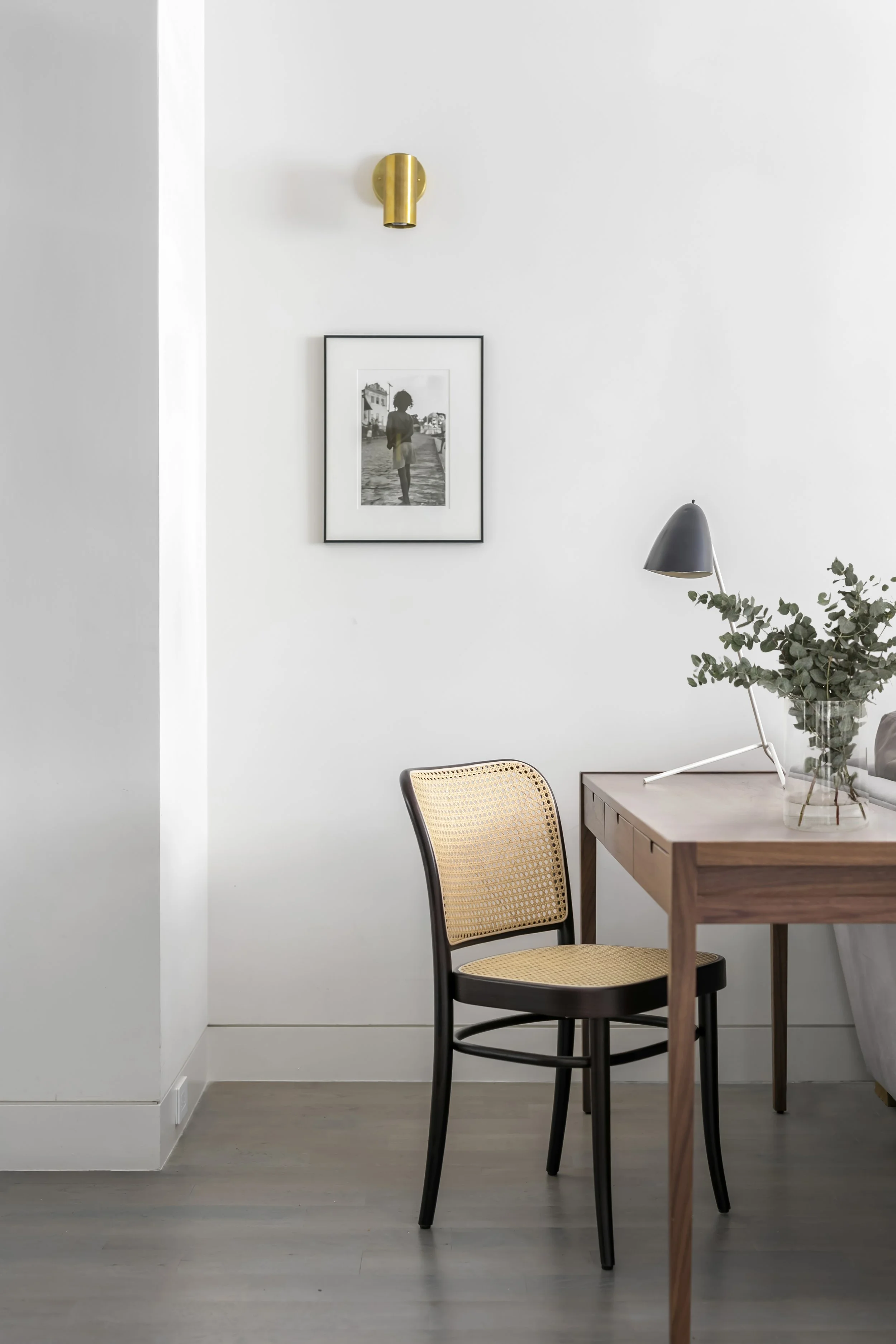 A minimalist interior scene featuring a black and tan chair with a caned backrest, a wooden desk, a black desk lamp, a glass vase with leafy green eucalyptus, a framed black-and-white photograph on the white wall, and a gold wall sconce.