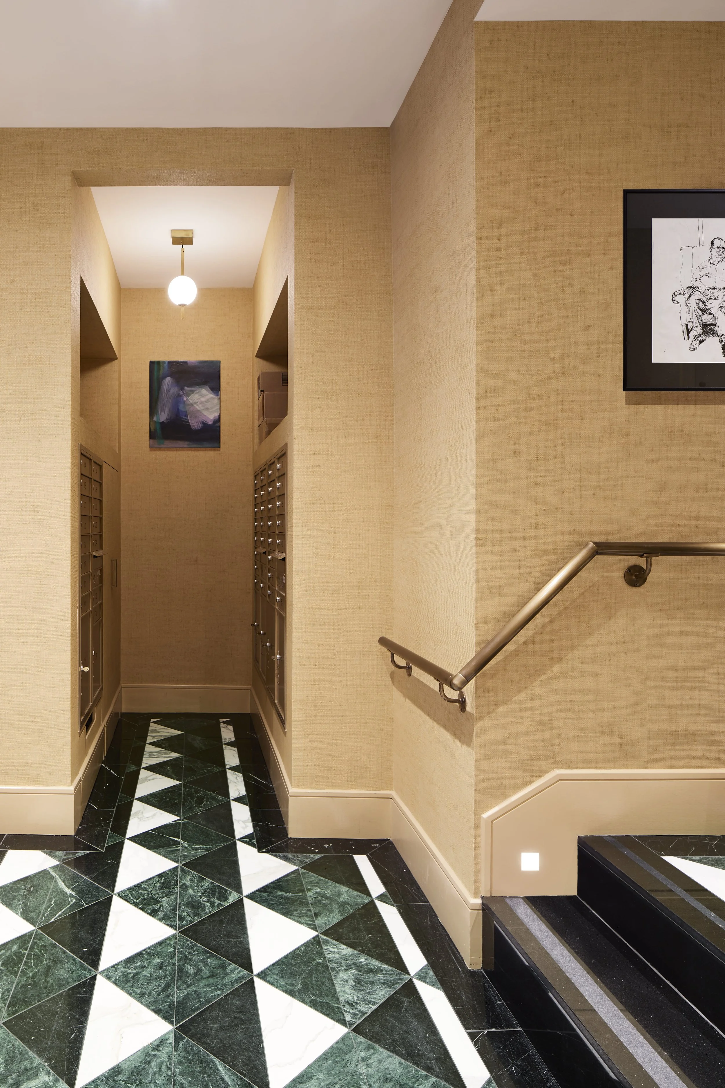 A hallway with beige walls, black and white geometric marble flooring, and a staircase with a metal handrail. Mailboxes are visible on the left side, and a small abstract picture hangs on the wall. There's a light fixture hanging from the ceiling.