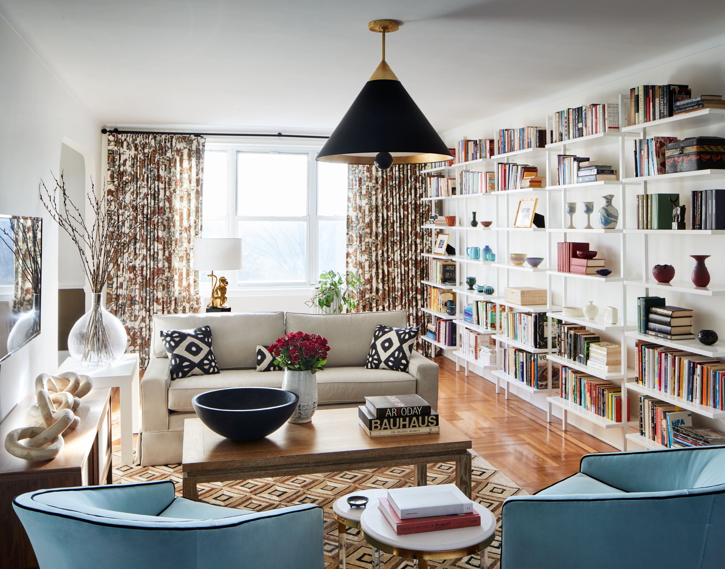 Bright living room with white walls, large window with patterned curtains, beige sofa with black and white pillows, blue armchairs, wooden coffee table with flower vase, floor lamp, white bookshelf filled with colorful books and decorative items, har