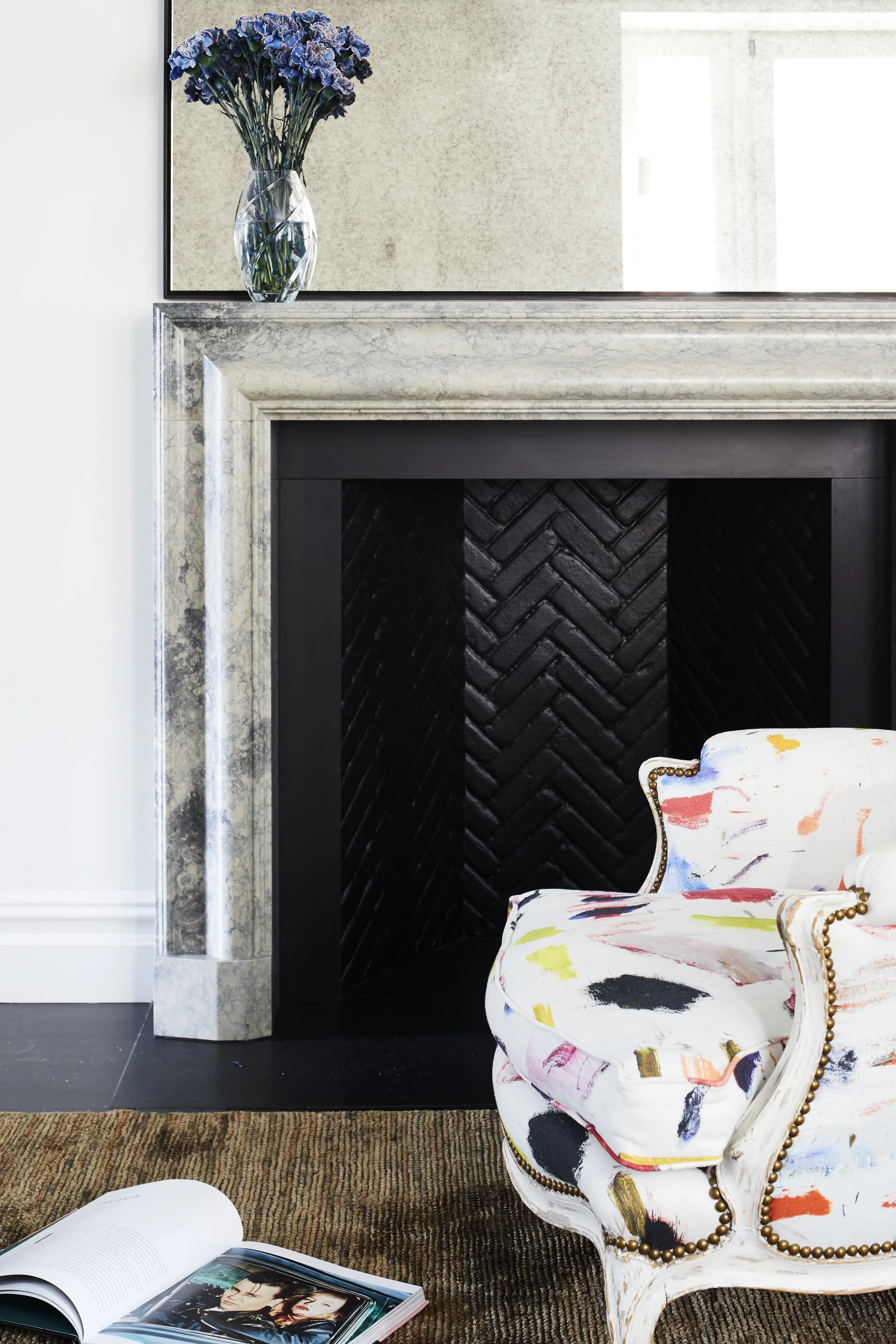 Close-up of a corner of a living room featuring a fireplace with a black textured interior, a marble mantel, a mirror above the mantel, a vase with purple flowers on the mantel, a colorful patterned armchair with a scalloped edge and gold nailhead tr
