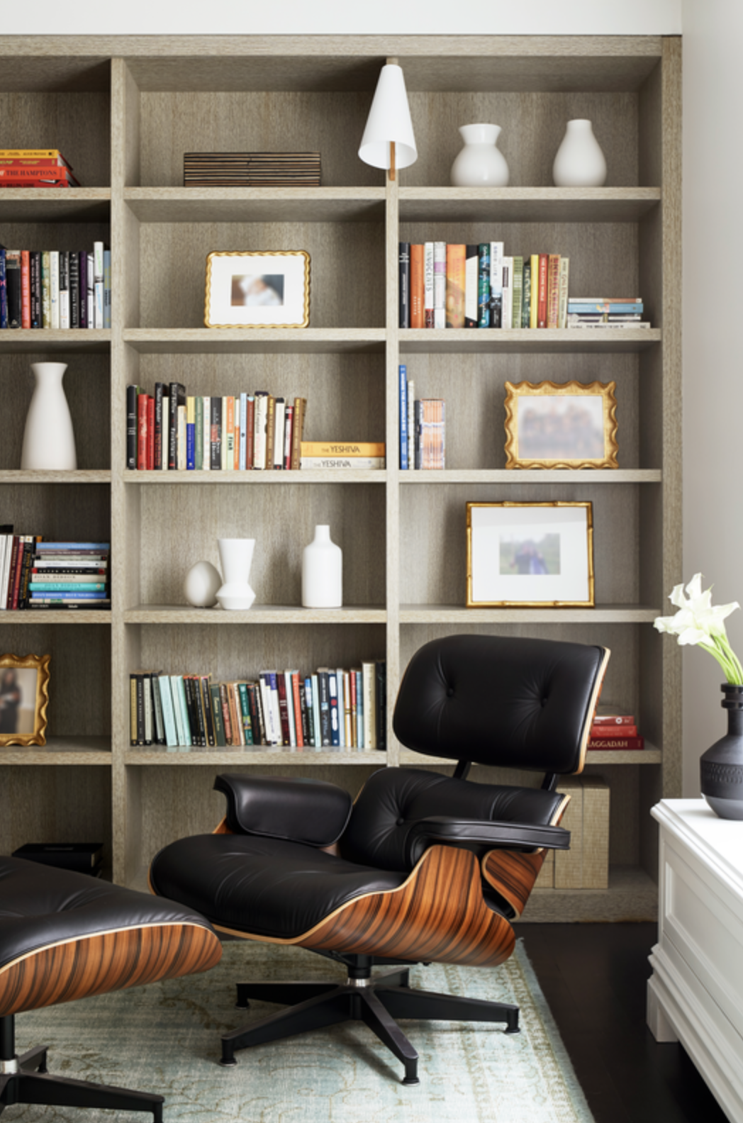 A room with a tall built-in bookshelf filled with books, white vases, and framed photos, a black leather and wood lounge chair, a white side table with a vase of white flowers, and a light-colored rug.