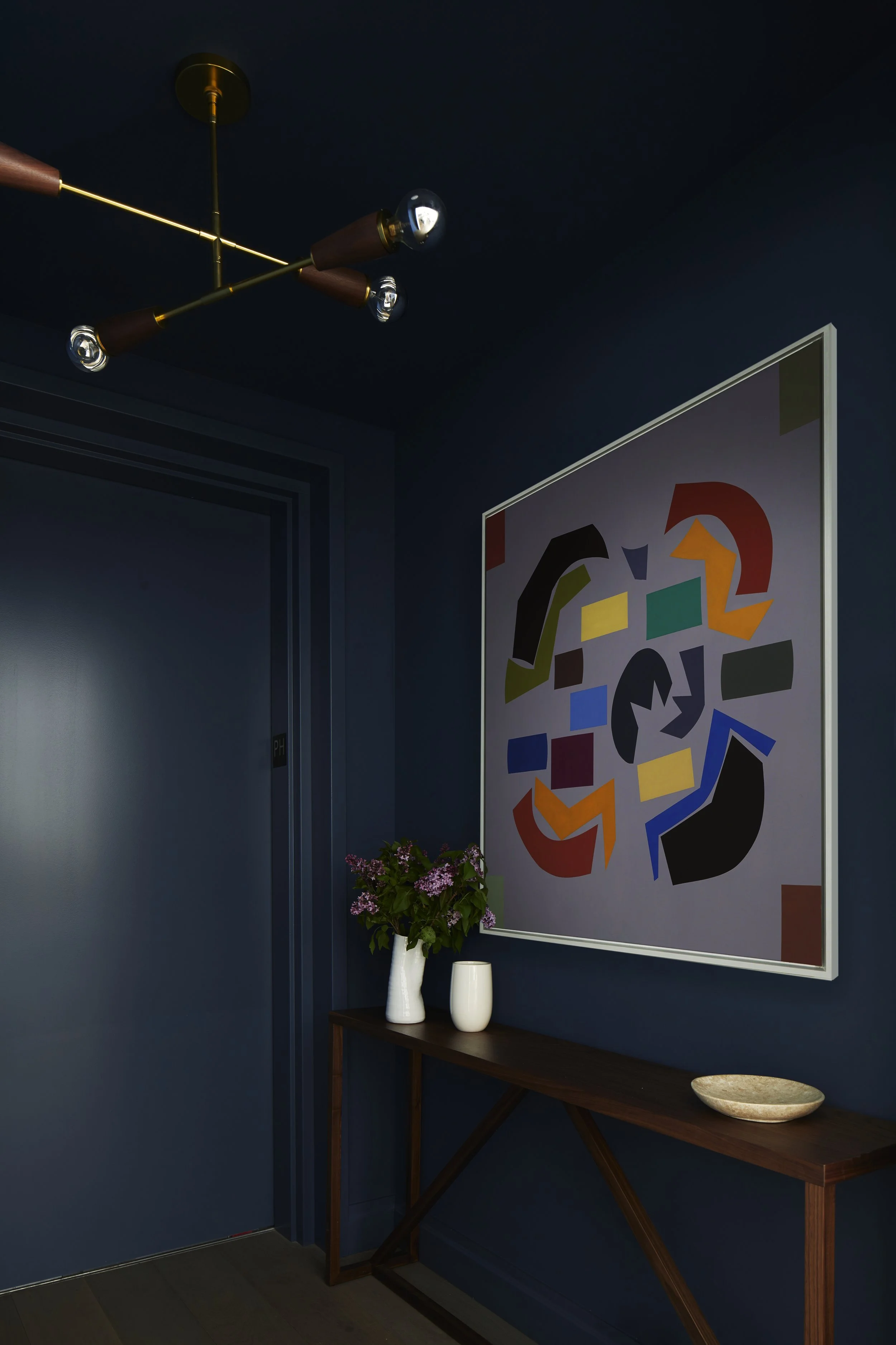 A corner of a room with a dark blue wall, a modern ceiling light fixture, a framed abstract art piece with colorful geometric shapes, a wooden console table with purple flowers in a white vase, a small white vase, and a beige decorative bowl.