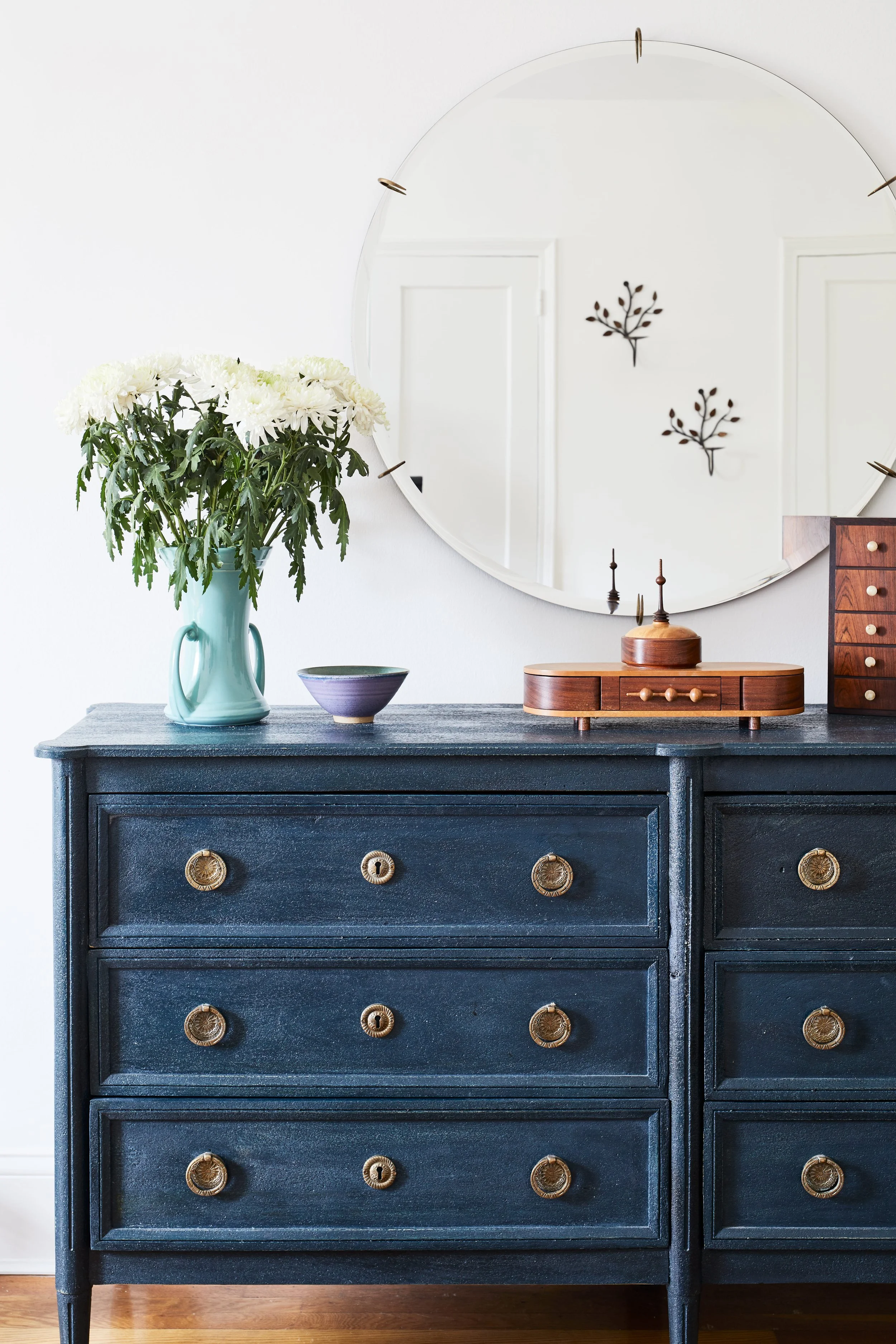 A dark blue vintage dresser with four drawers and gold knobs, decorated with a turquoise vase of white flowers, a small purple bowl, and a wooden jewelry box. A round mirror hangs above, reflecting two black metal wall art pieces resembling branches.