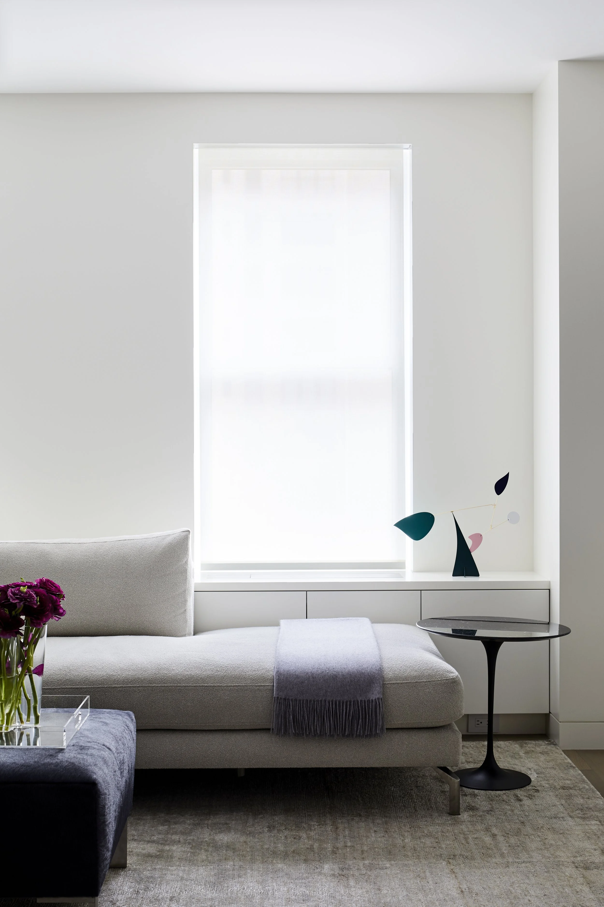 Modern living room with a large window, a light-colored sofa with a gray throw blanket, a black side table, a vase of purple flowers, and minimalist wall art.