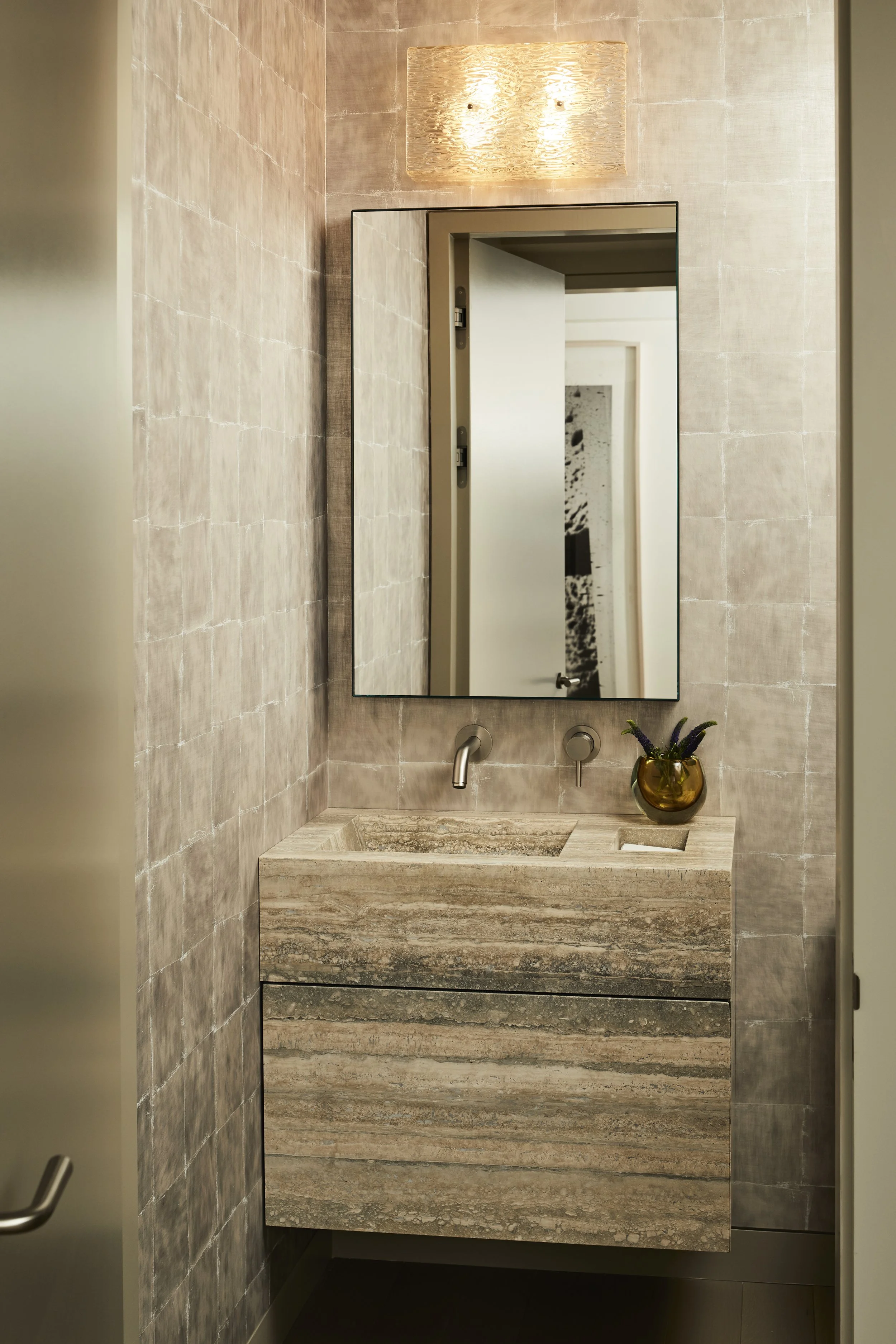 A modern bathroom with a beige textured stone sink, round wall-mounted faucet, and a rectangular mirror above. There is a brass-colored vase with purple flowers on the right side of the sink, and the wall and countertop are made of matching beige sto