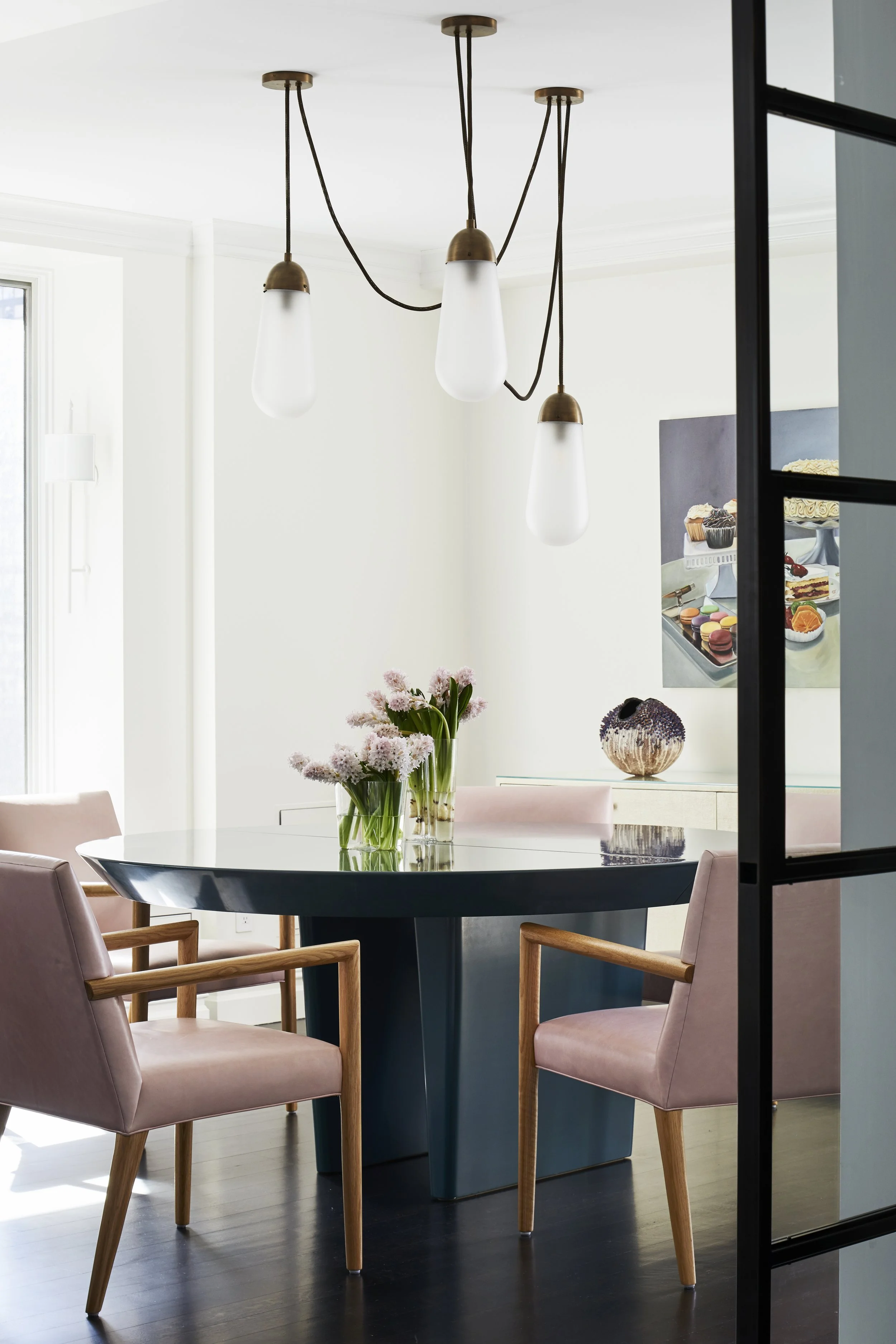A modern dining room with a round black table, pink upholstered chairs with wooden armrests, a glass vase with pink flowers, a white sideboard, and a chandelier with three hanging white glass pendant lights.