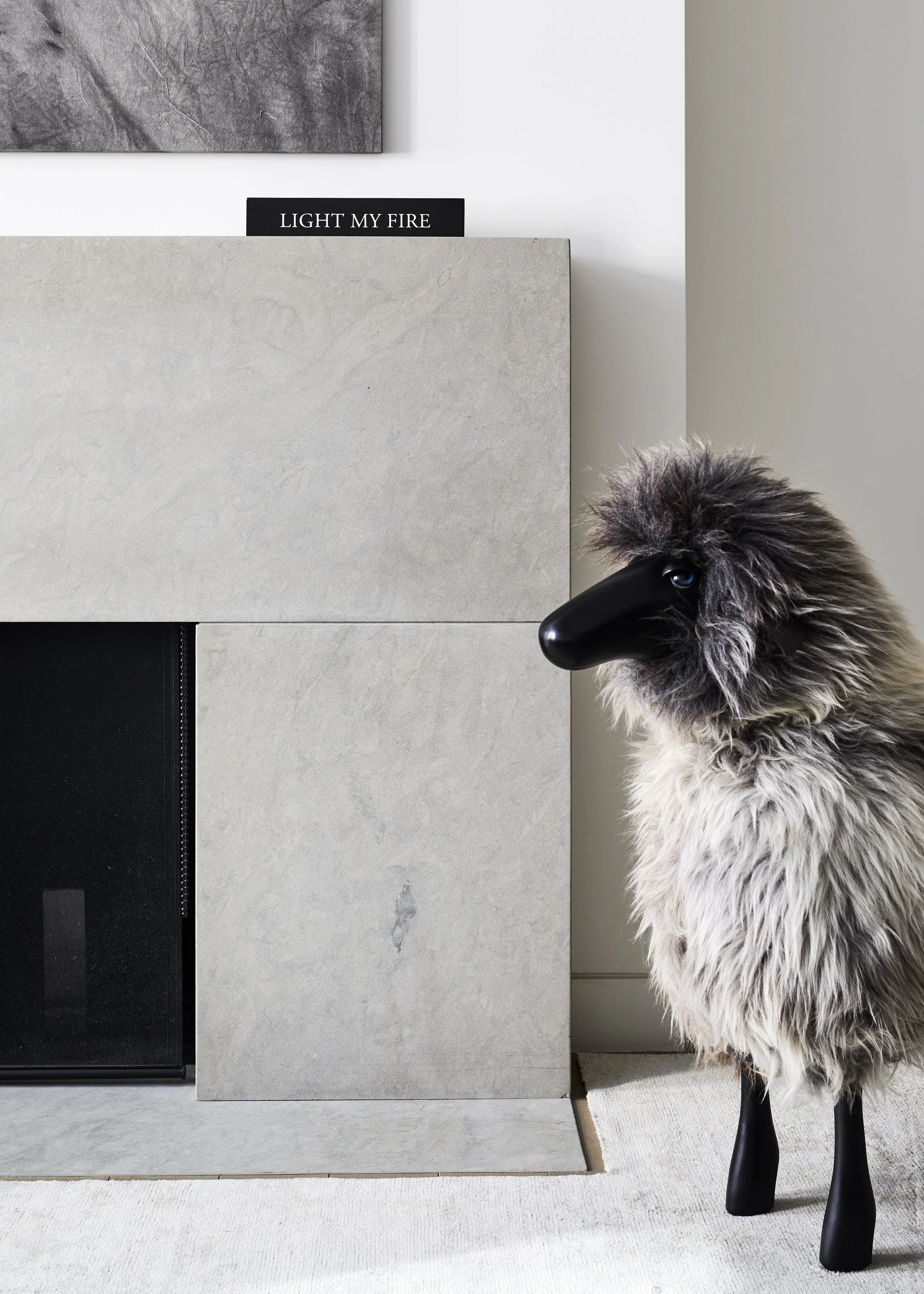 A decorative sheep sculpture with a black face and wooden legs positioned next to a modern fireplace with a concrete surround. There is a black sign on the wall behind it that says "LIGHT MY FIRE."