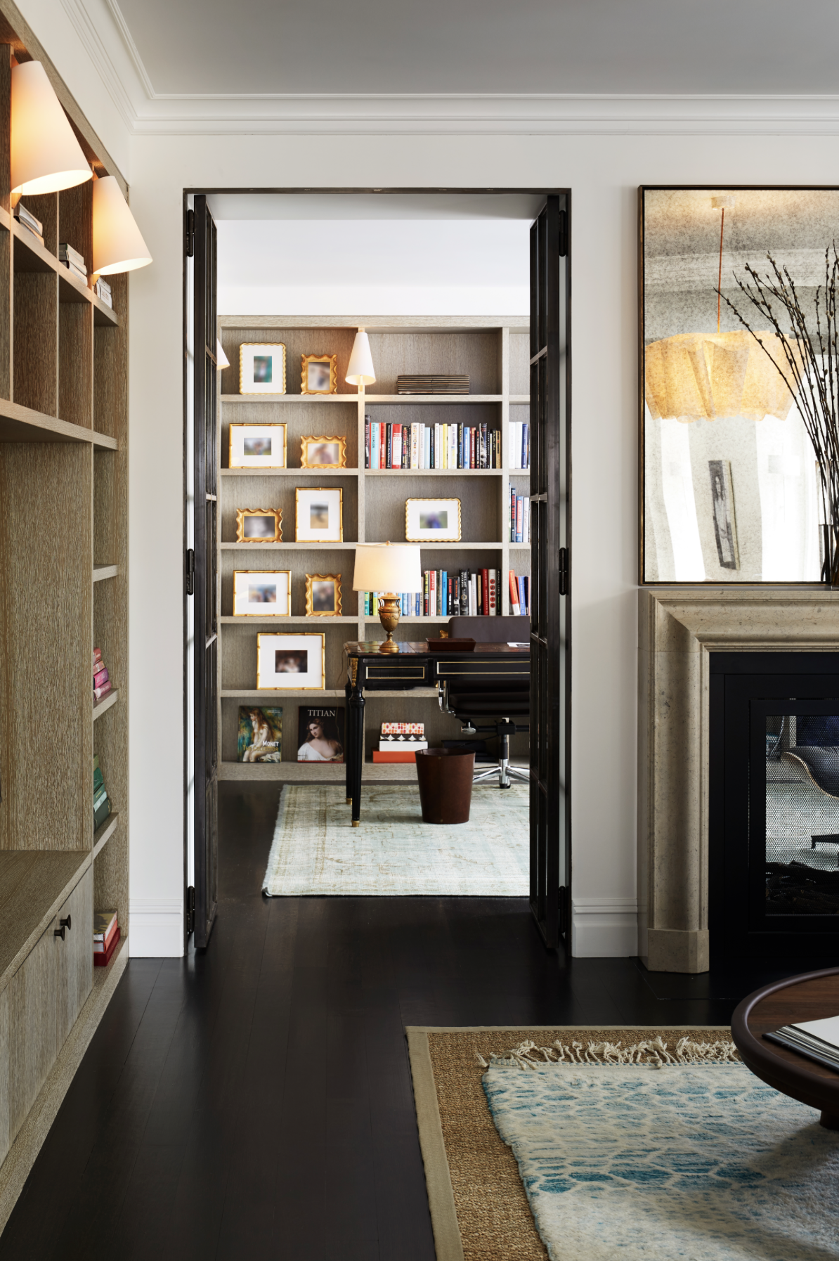 A cozy home office with a black desk, a chair, a table lamp, and a bookcase filled with books and photo frames, viewed through an open doorway from a living room with a fireplace and a patterned rug.