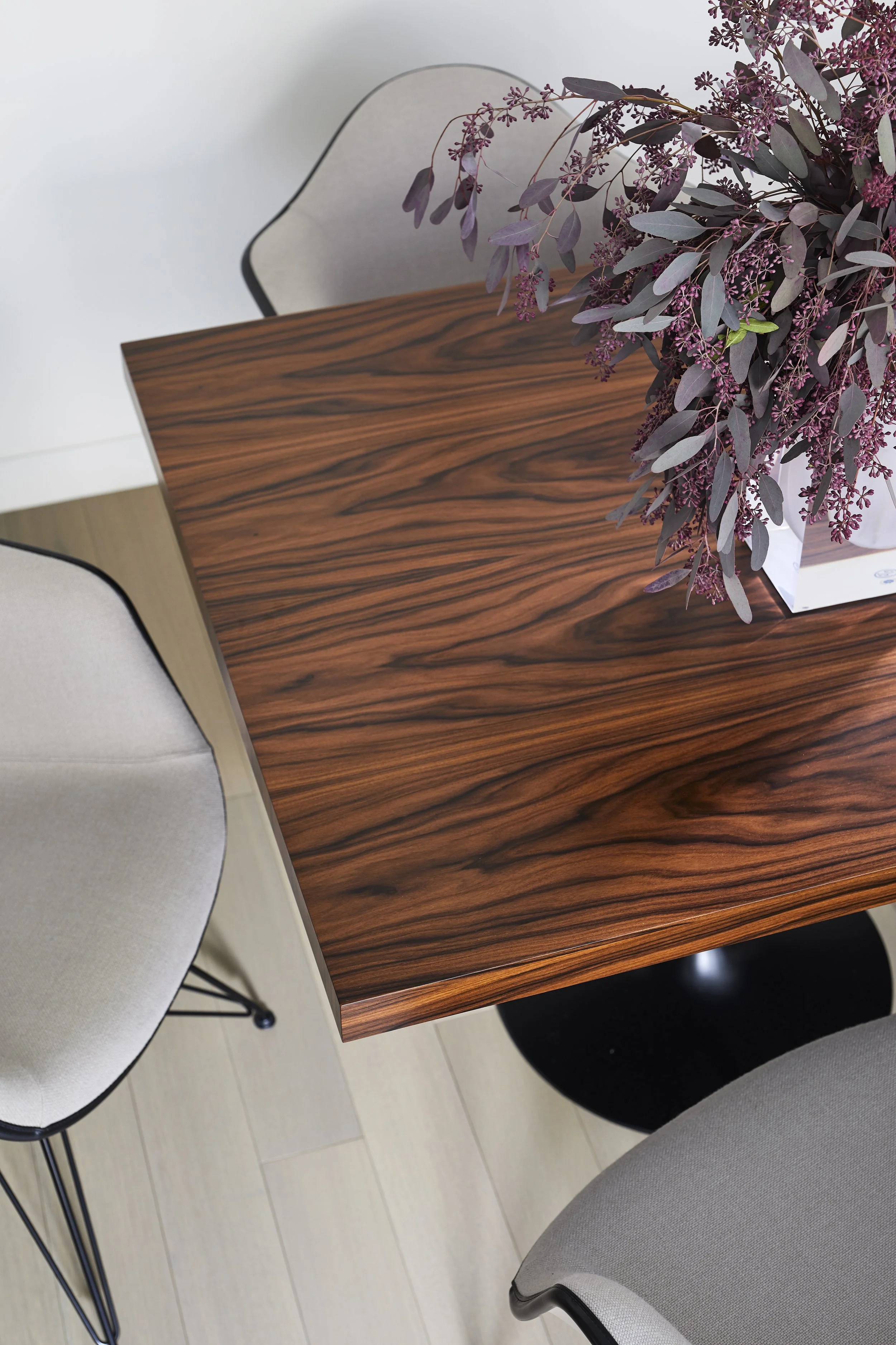 A wooden dining table with a floral arrangement in a white box and two upholstered chairs with black trim, one beige and one cream.