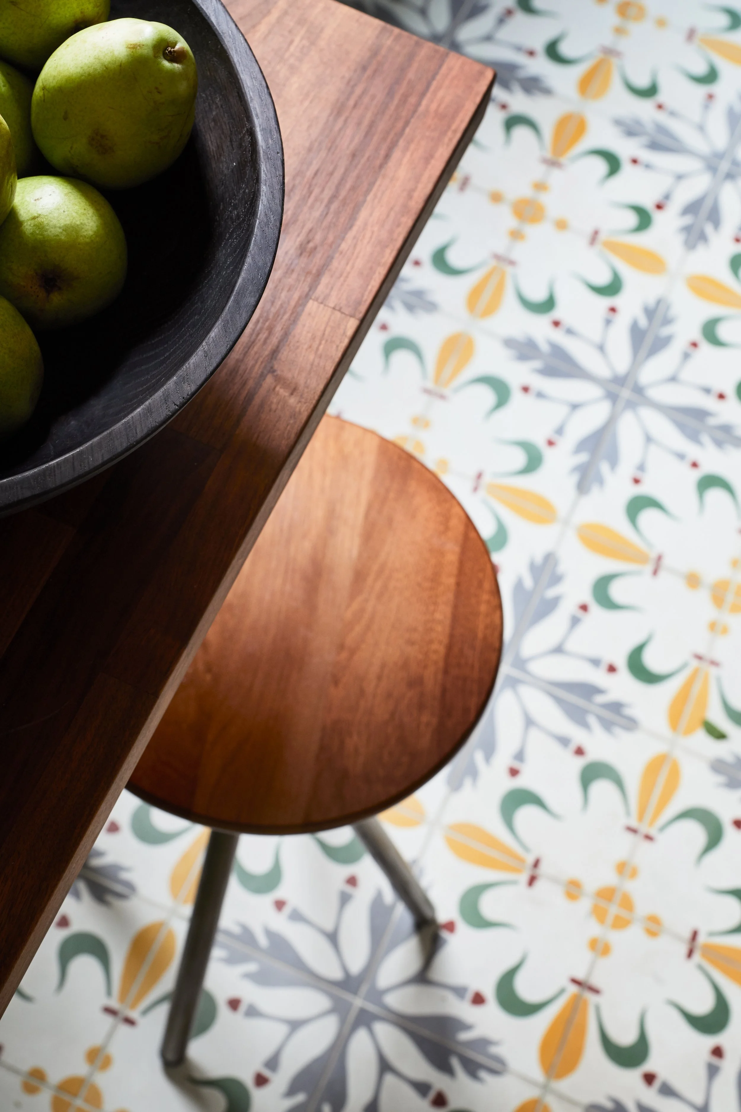A top-down view of a wooden table with a black bowl containing green apples, and a wooden chair on patterned tile flooring with floral designs.