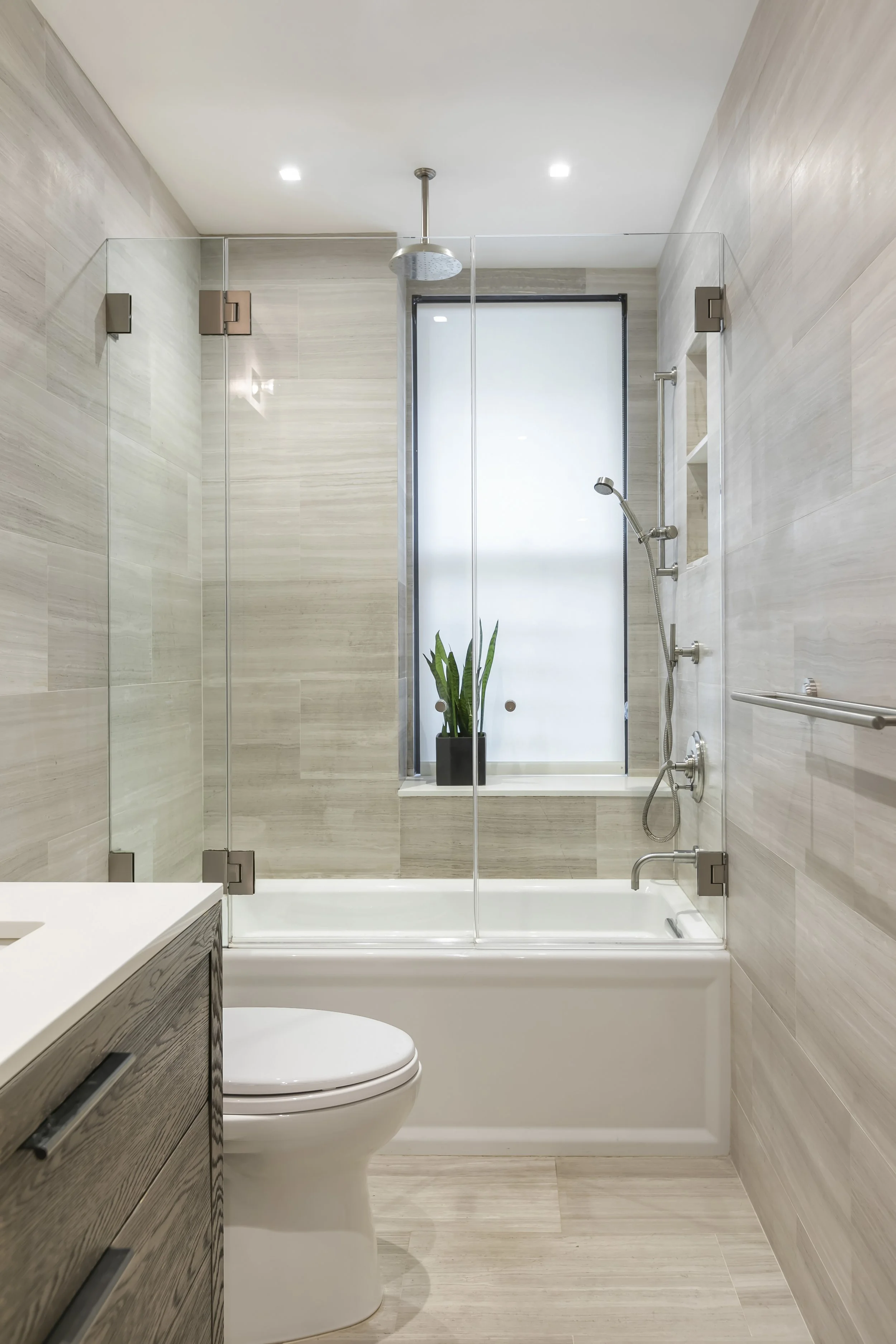 Modern bathroom with glass shower enclosure, bathtub, white toilet, vanity with dark wood drawers, gray wood-like tiled walls, a large frosted window, and a potted plant.