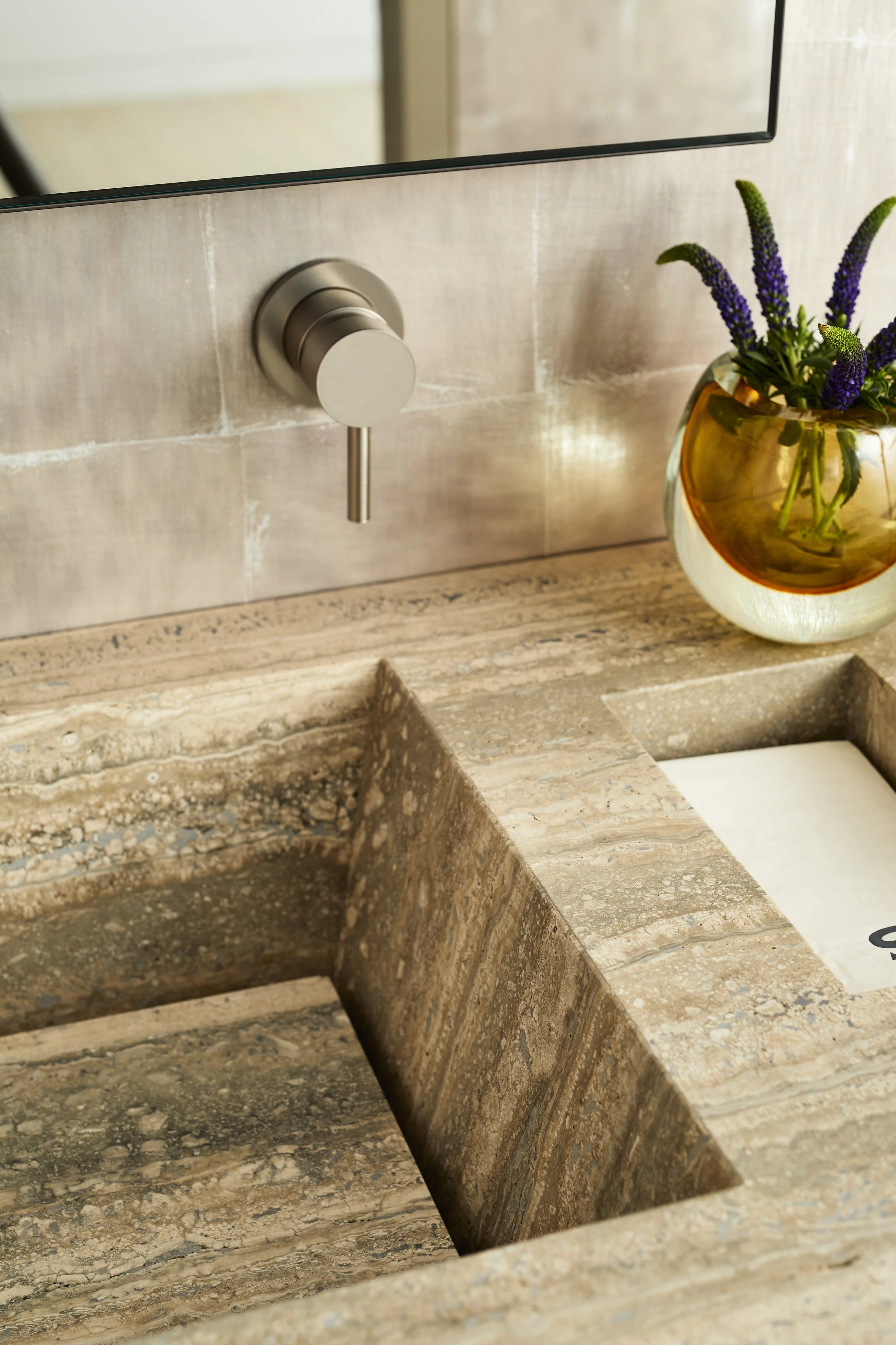 Close-up of a beige marble sink with a modern metallic faucet and a vase with purple flowers on the countertop.