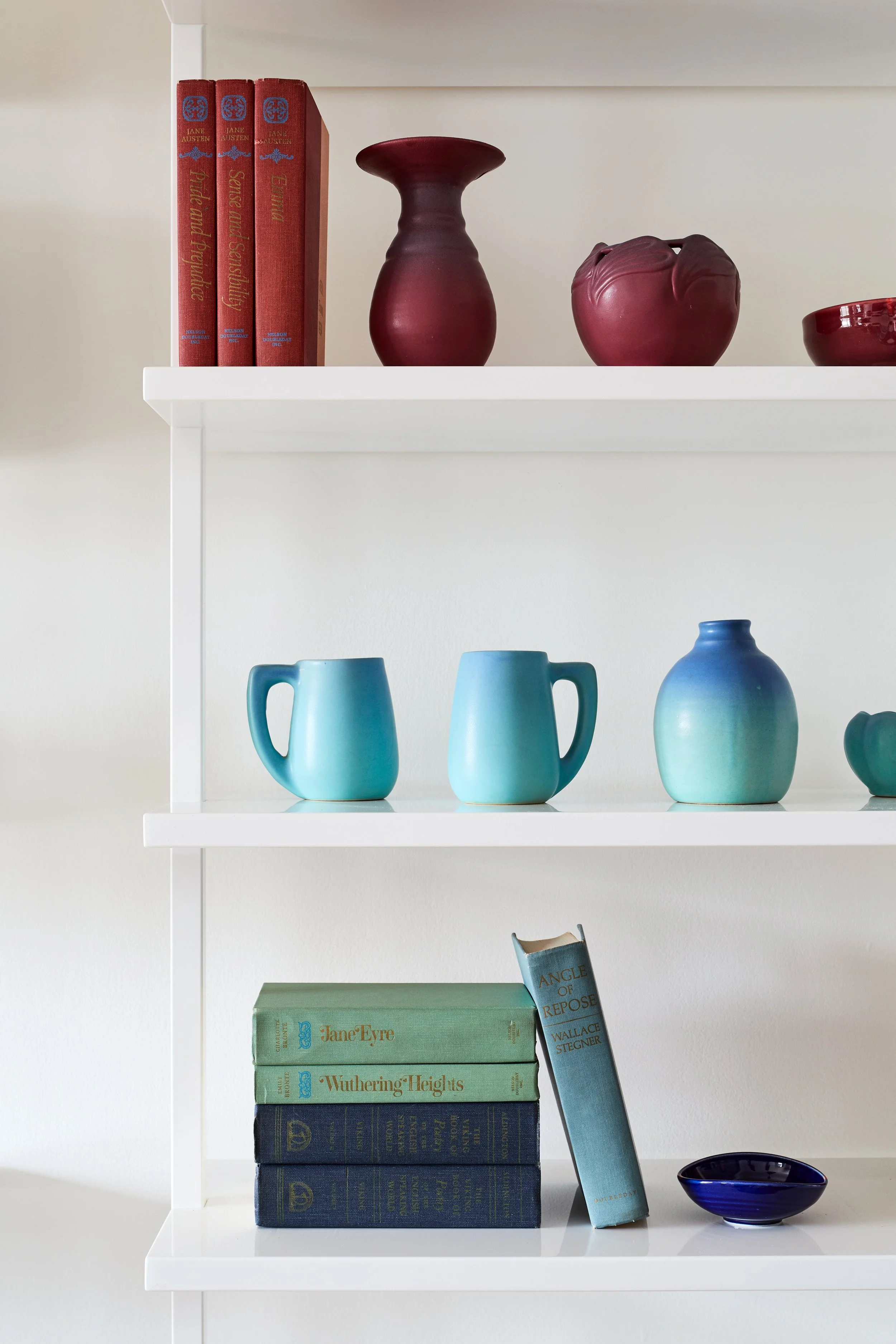 White bookshelf with red and blue books, burgundy vases, blue mugs, and a small blue bowl.