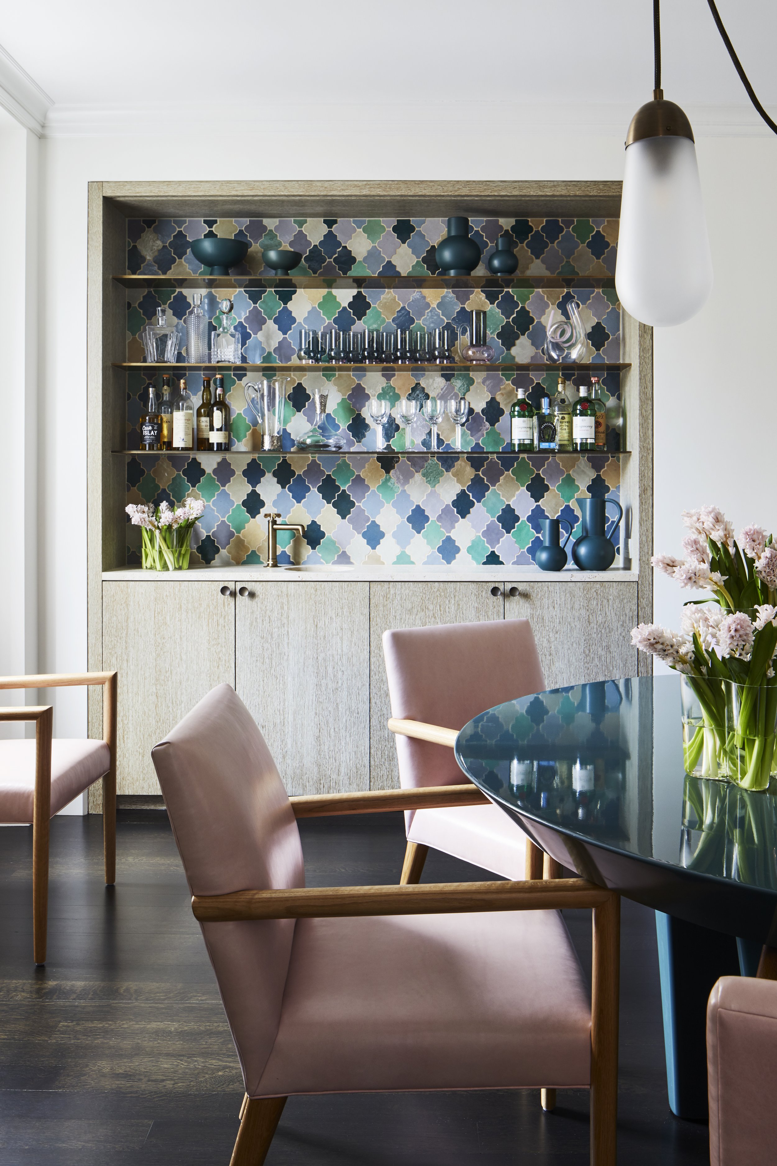 A dining room with a built-in bar featuring colorful patterned tiles, glassware, liquor bottles, and blue vases, with pink chairs and a vase of pink flowers on a dark table.