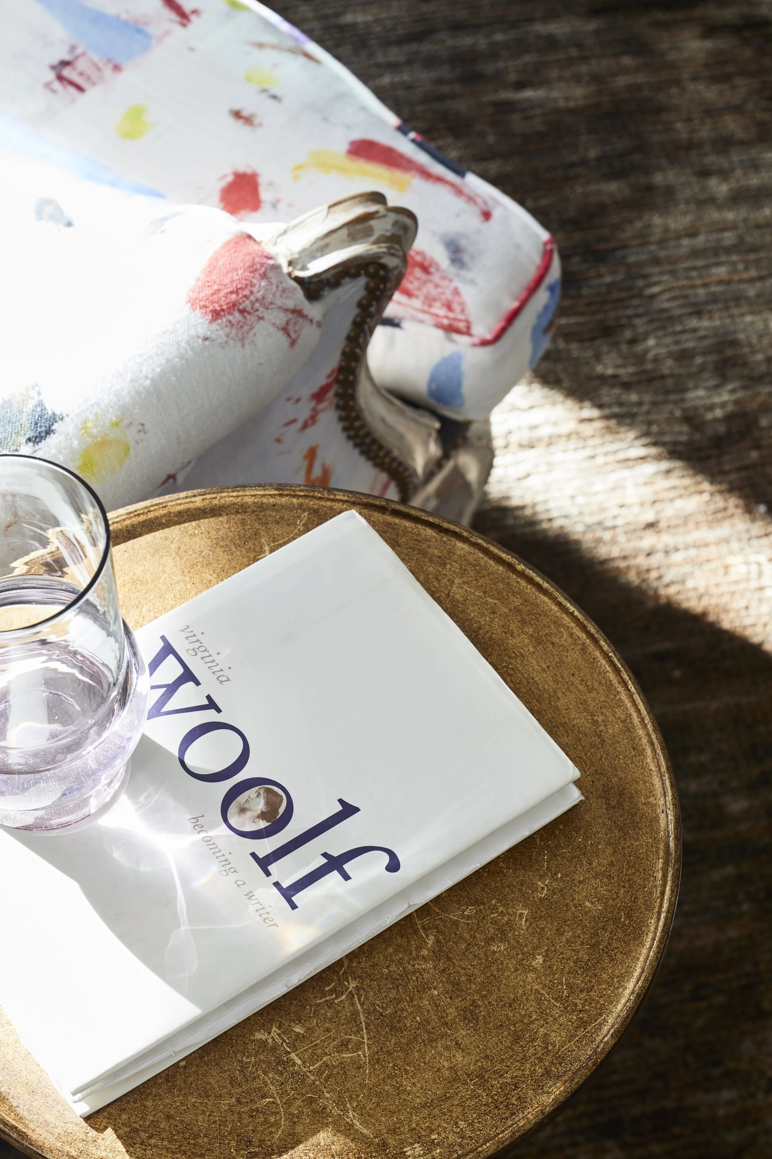 A close-up of a circular wooden table with a book titled 'VOIF' by Virginia on it, a glass of water, and sunlight casting shadows. In the background, there's a colorful, painted, vintage-looking sofa.