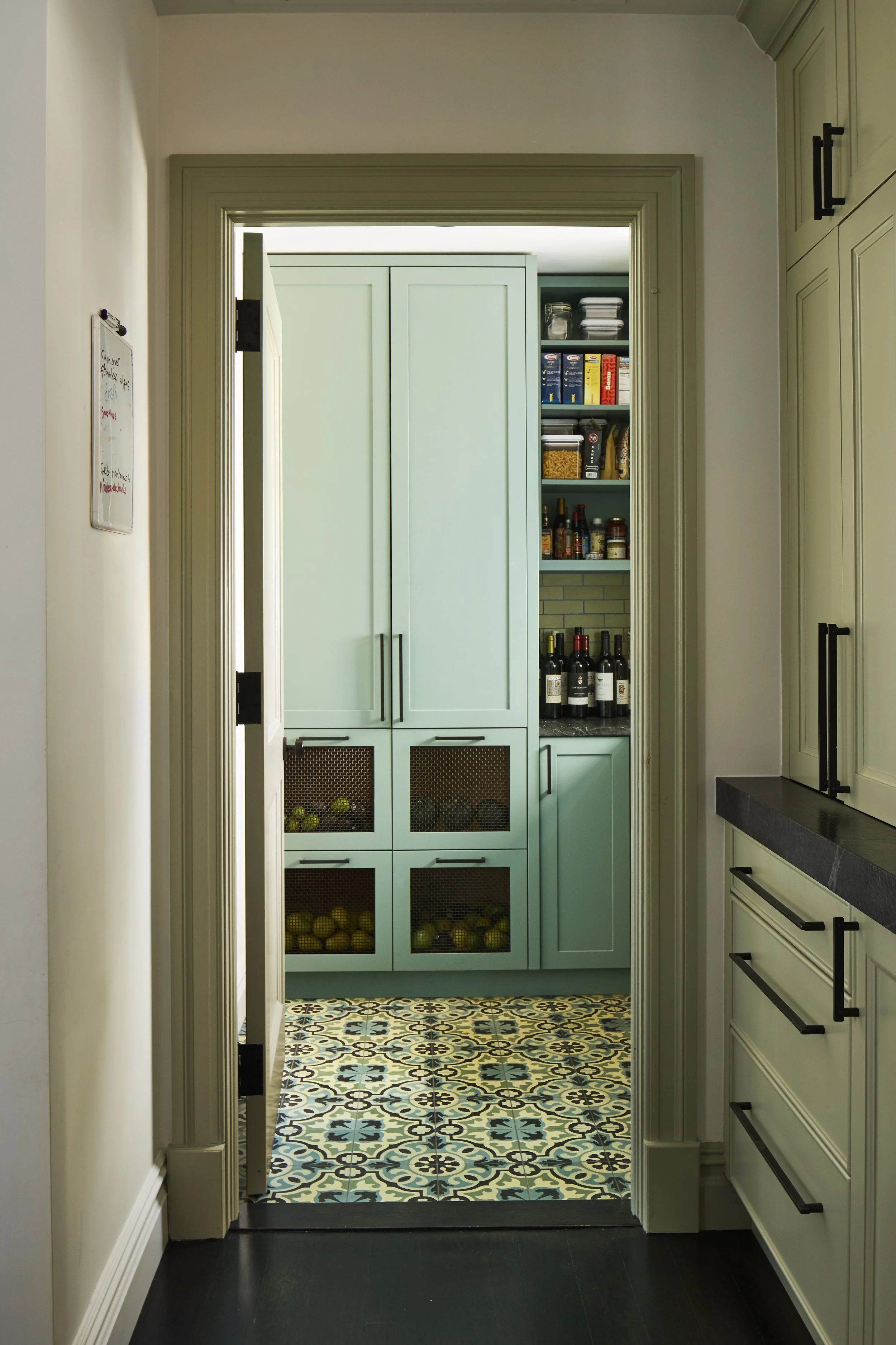 View into a pantry area through an open door, showing cabinets, shelves with food and bottles, and a decorative patterned tile floor.