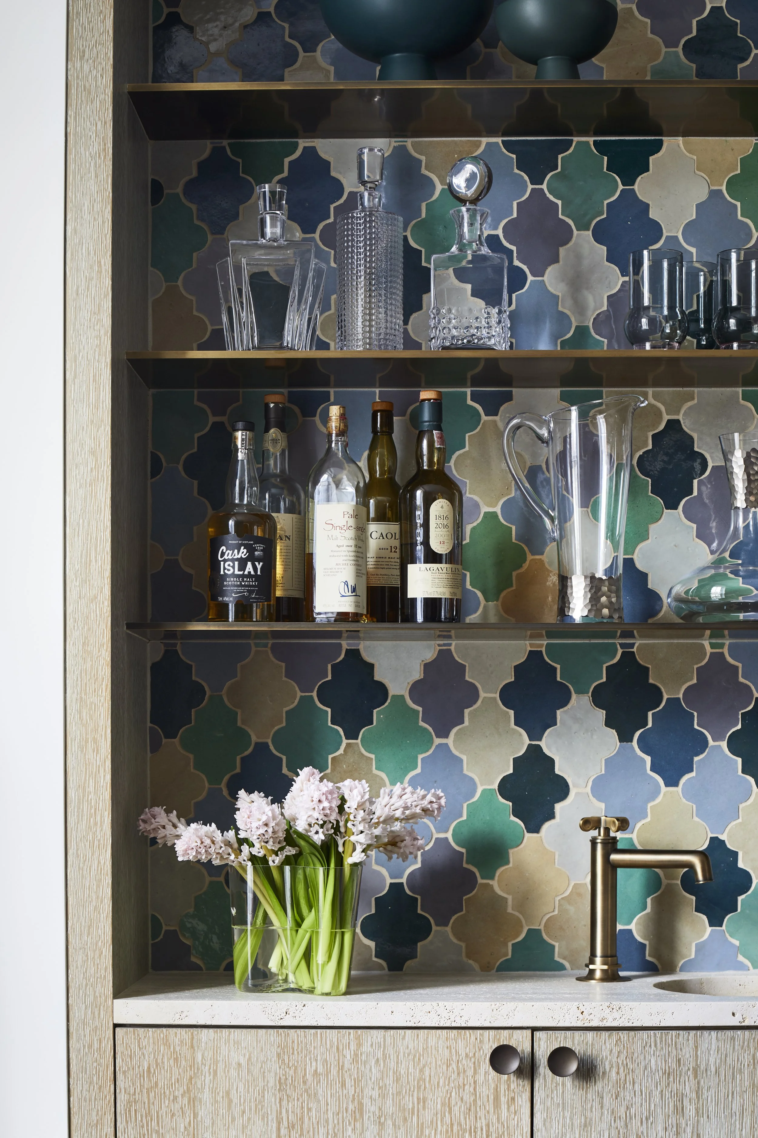 A bar cabinet with glass bottles, decanters, and glasses on glass shelves, a brass faucet, a vase with pink flowers, and a tiled wall with blue, green, and beige patterns.