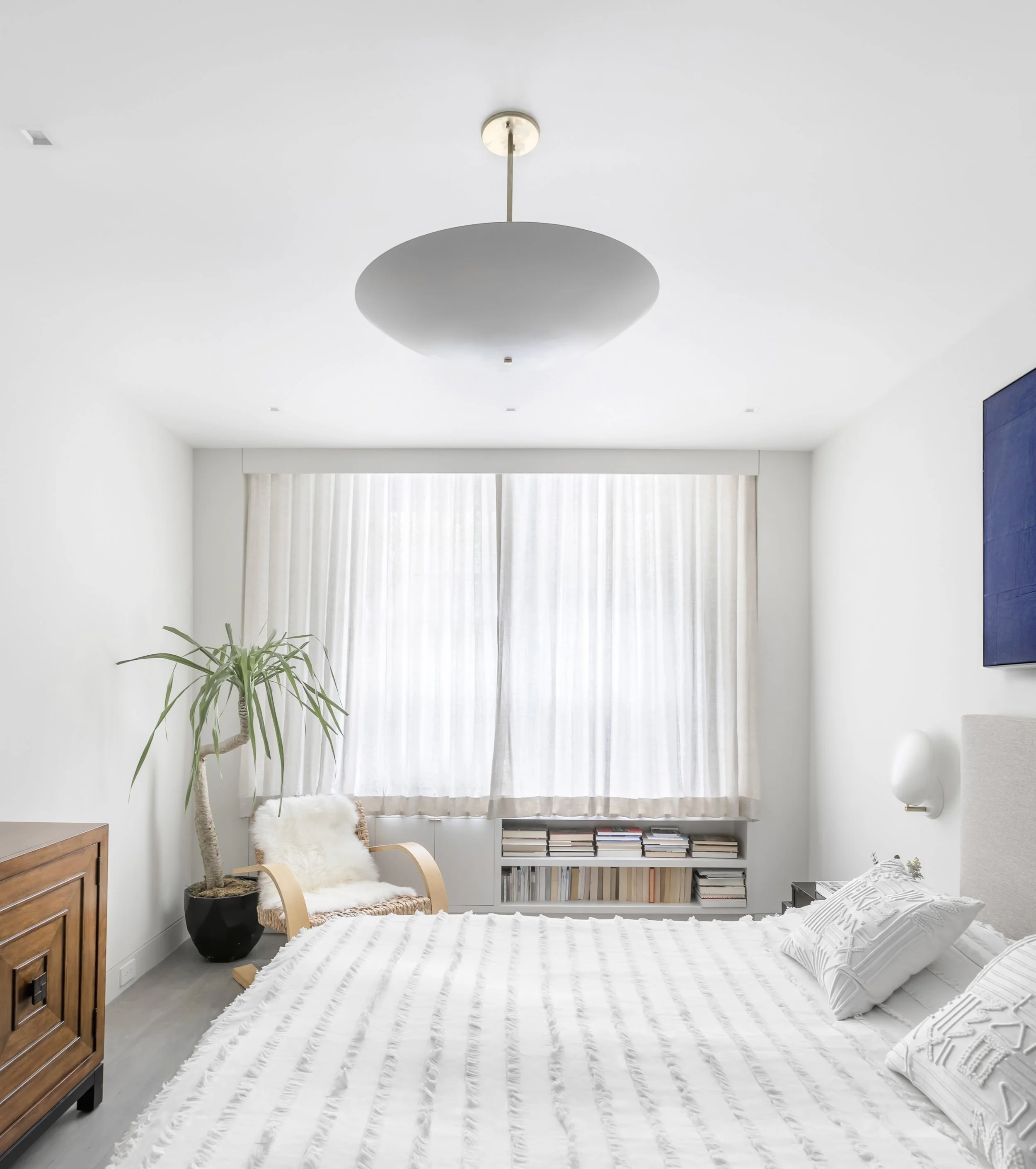 Minimalist bedroom with white walls, a large bed with white textured bedding and pillows, a wooden nightstand, a large plant in a black pot, a reading chair with a fur cushion, and a window with white curtains.